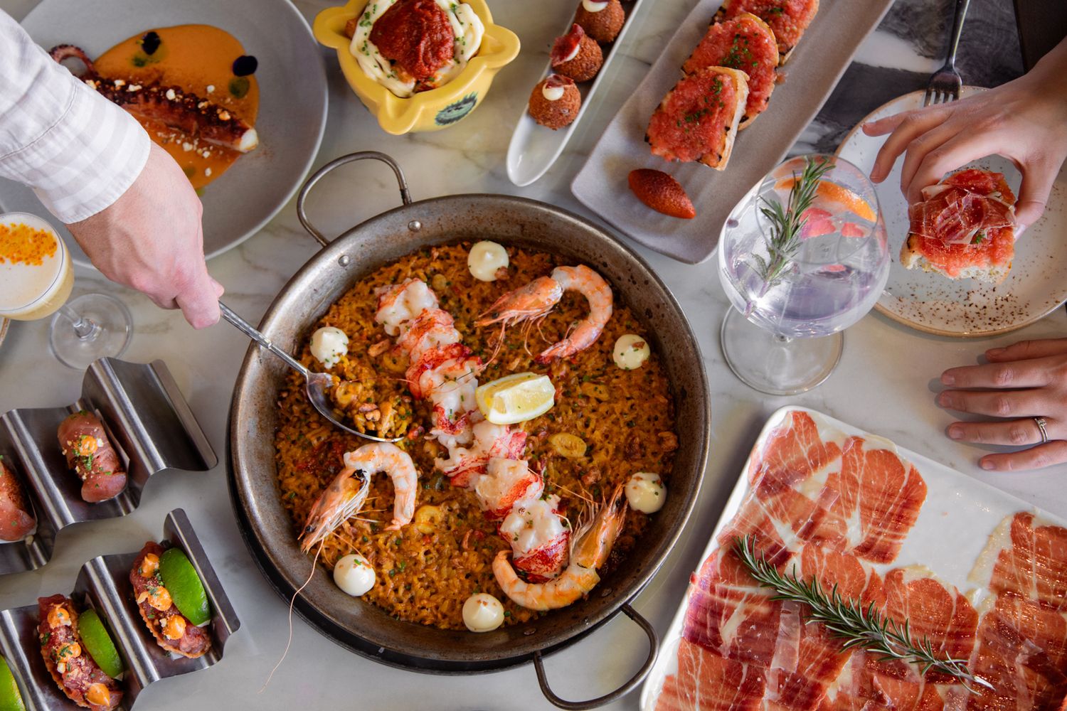 Overhead of spanish tapas dishes, cocktails, and seafood paella in the center. Hands reach in from both sides to enjoy food