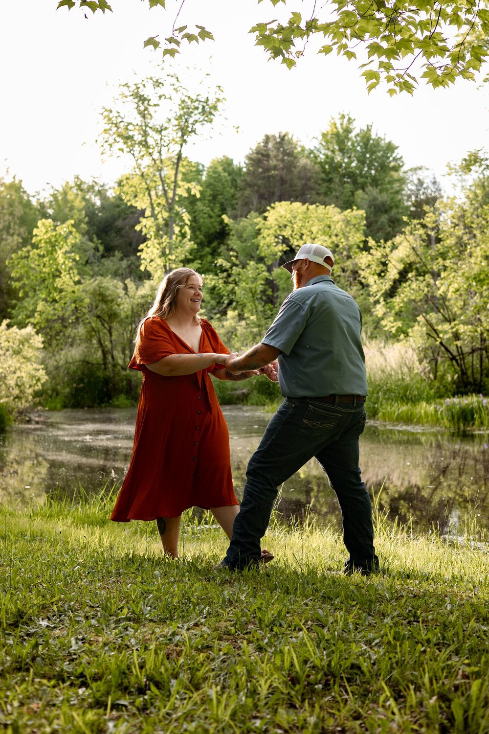 Couple dancing and holding hands in a lush green park by a pond, woman in red dress smiling at man in blue shirt.