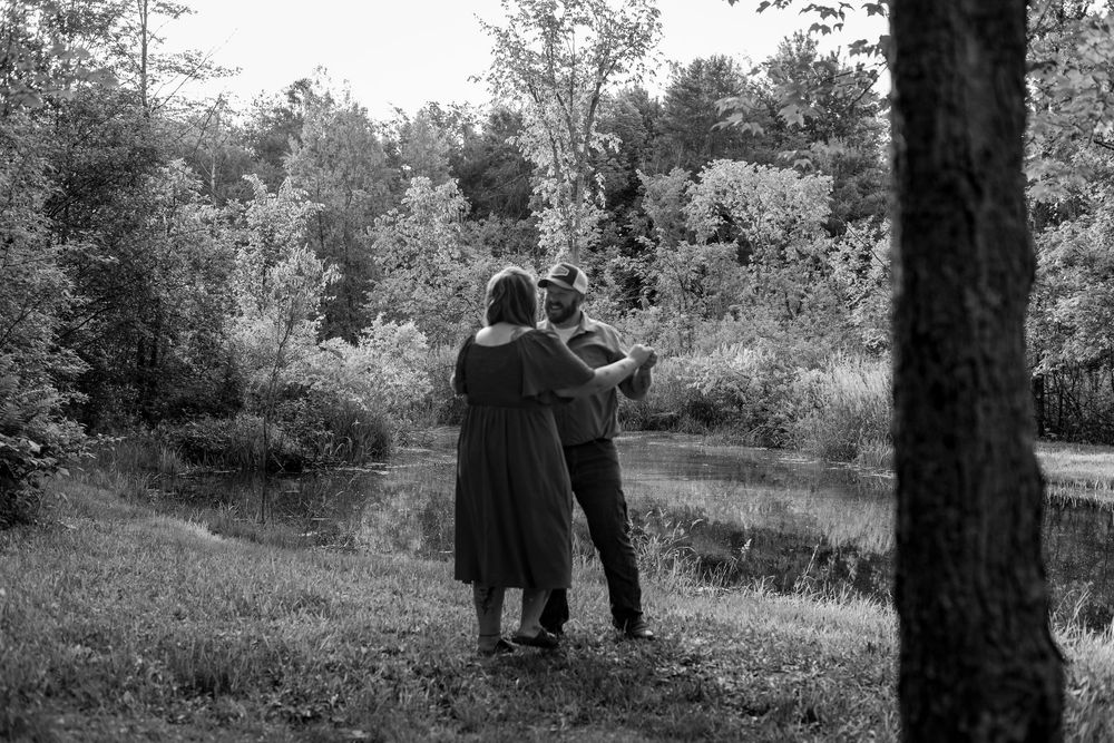 Black and white photo of a couple standing together near a serene pond surrounded by lush trees.