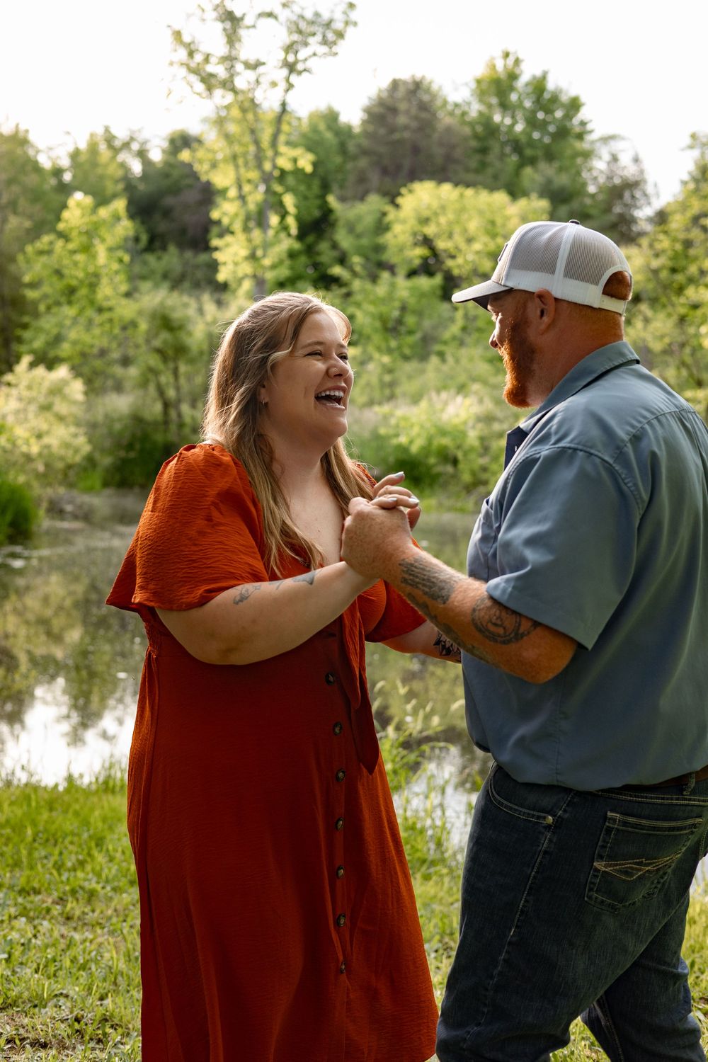 Laughing couple holding hands outdoors near a pond, woman in red dress and tattooed man in blue shirt and white cap.
