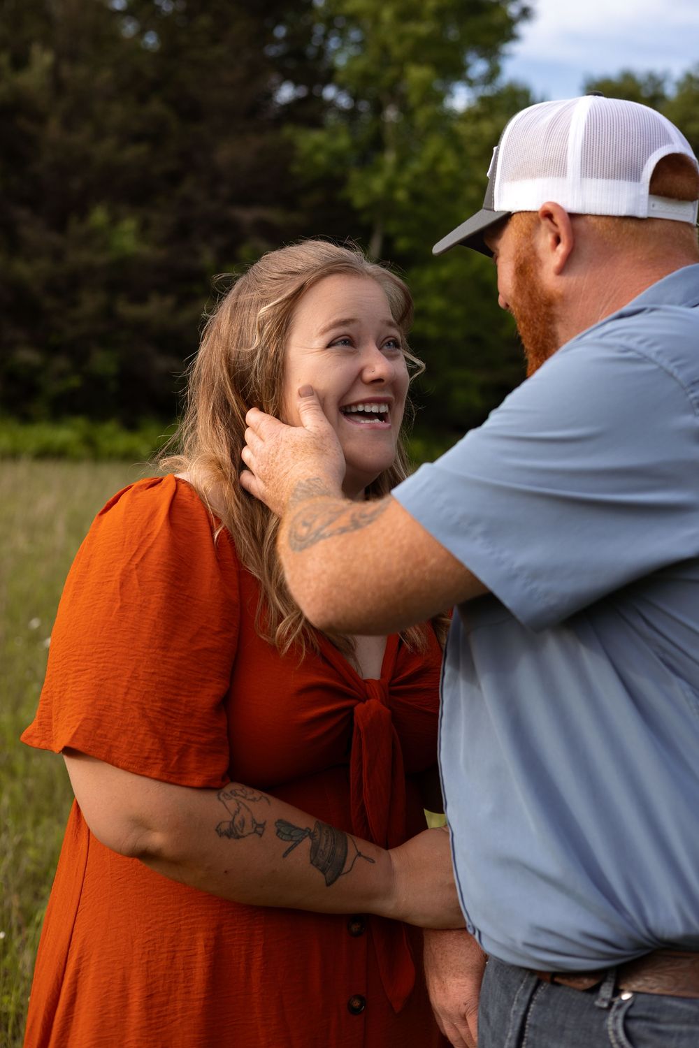 A laughing woman in an orange dress and tattooed man in a blue shirt share a tender moment in a green field.