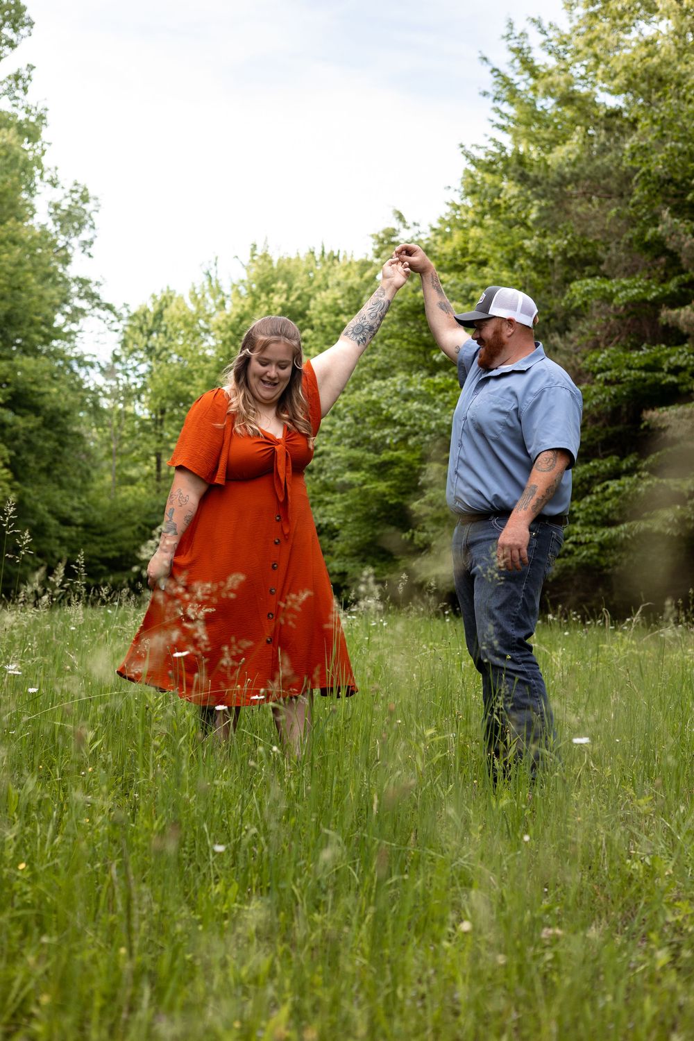 A couple dances joyfully in a lush green meadow, woman in red dress twirling as man in blue shirt holds her hand.