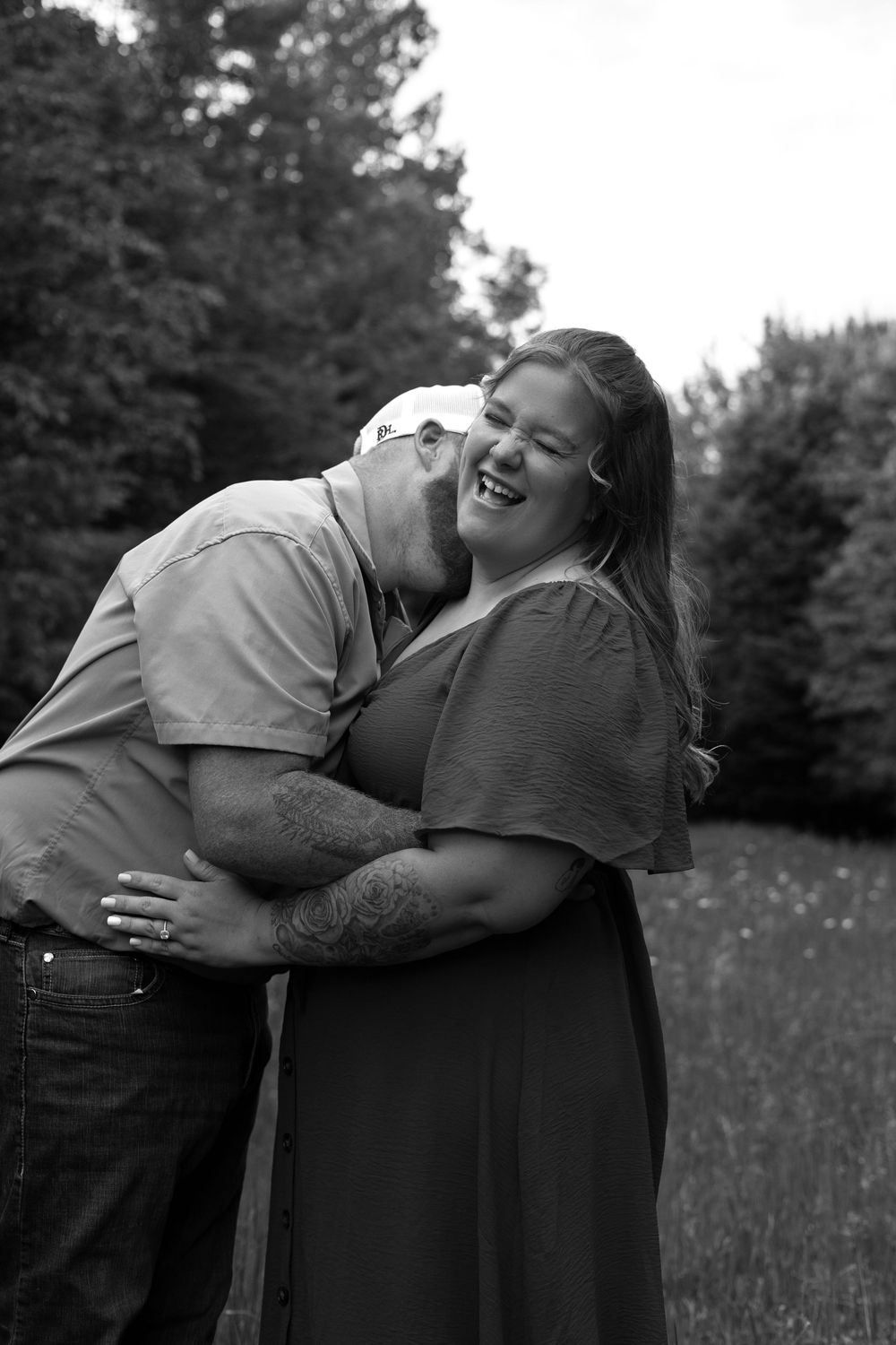 Black and white photo of a couple embracing outdoors, man kissing womans cheek as she laughs joyfully.