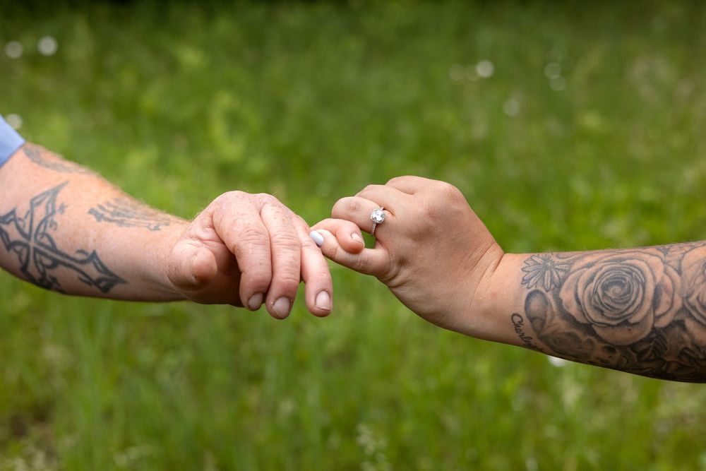 Two tattooed hands gently touching fingers outdoors, with one hand wearing a diamond engagement ring.