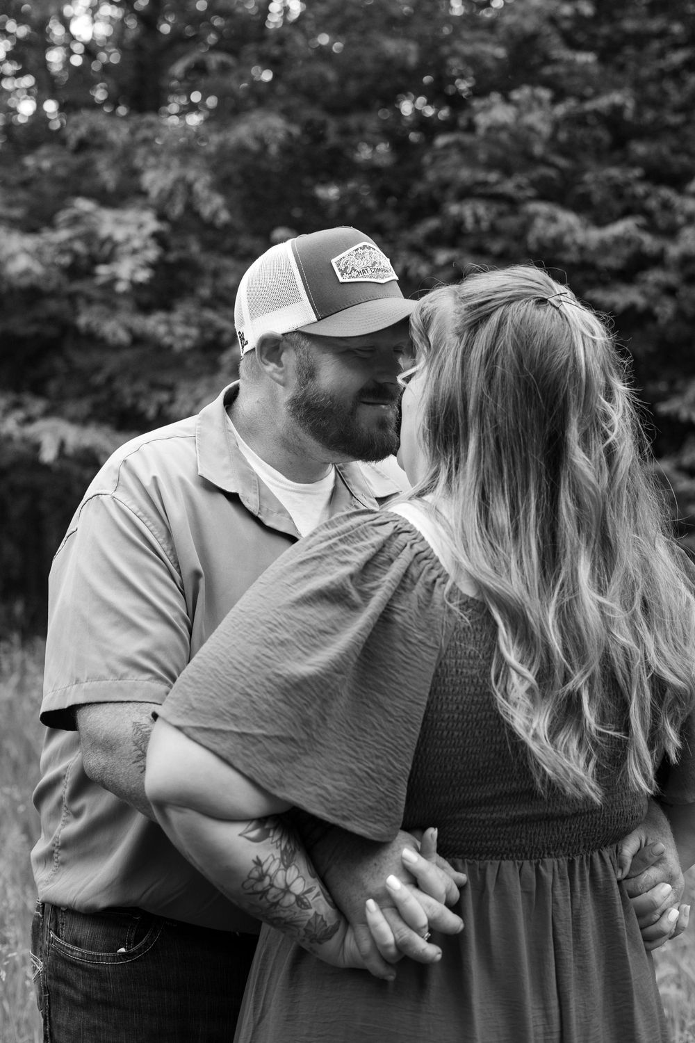 Black and white photo of a bearded man in a trucker hat embracing a woman with long hair outdoors.