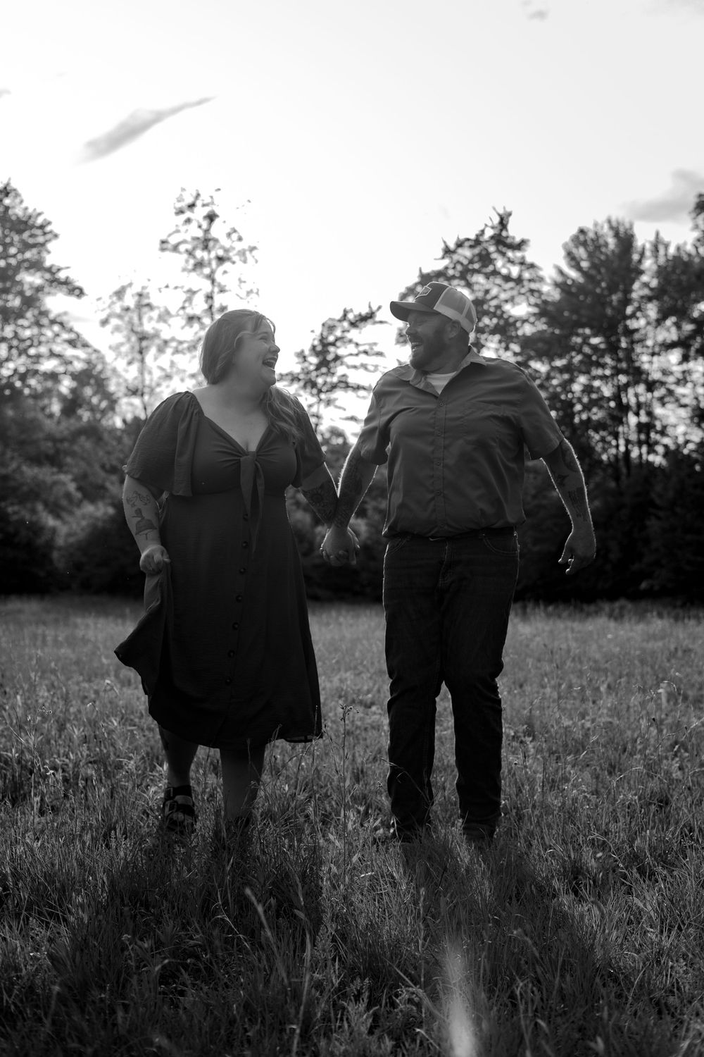 Black and white photo of a couple holding hands and walking through a grassy field with trees in the background.