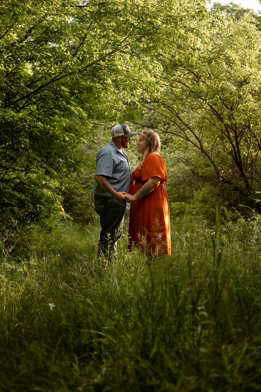 Couple holding hands in a lush green forest clearing, woman in orange dress and man in blue shirt and cap.