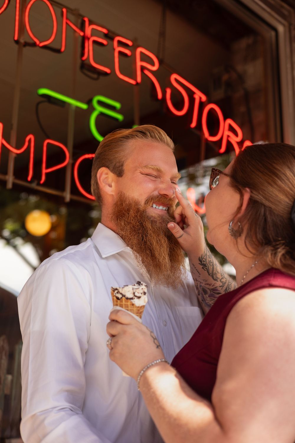 Couple laughing and sharing ice cream cone outside Pioneer Store with neon signs, man with red beard in white shirt.