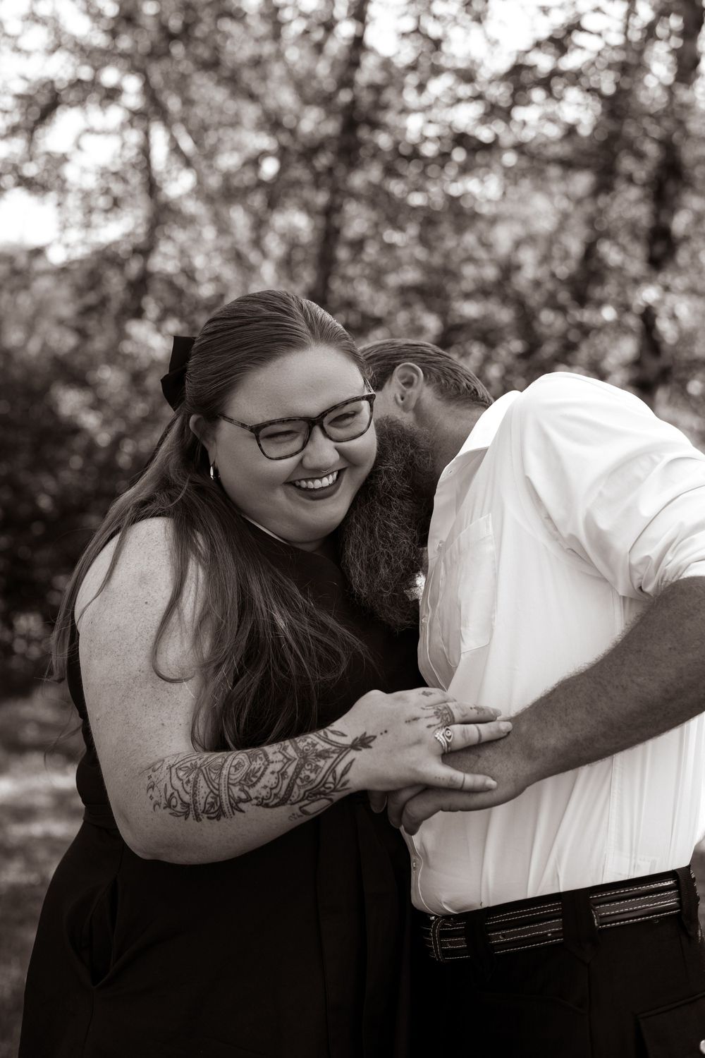Black and white photo of a smiling woman with glasses and tattoos embracing a man outdoors.
