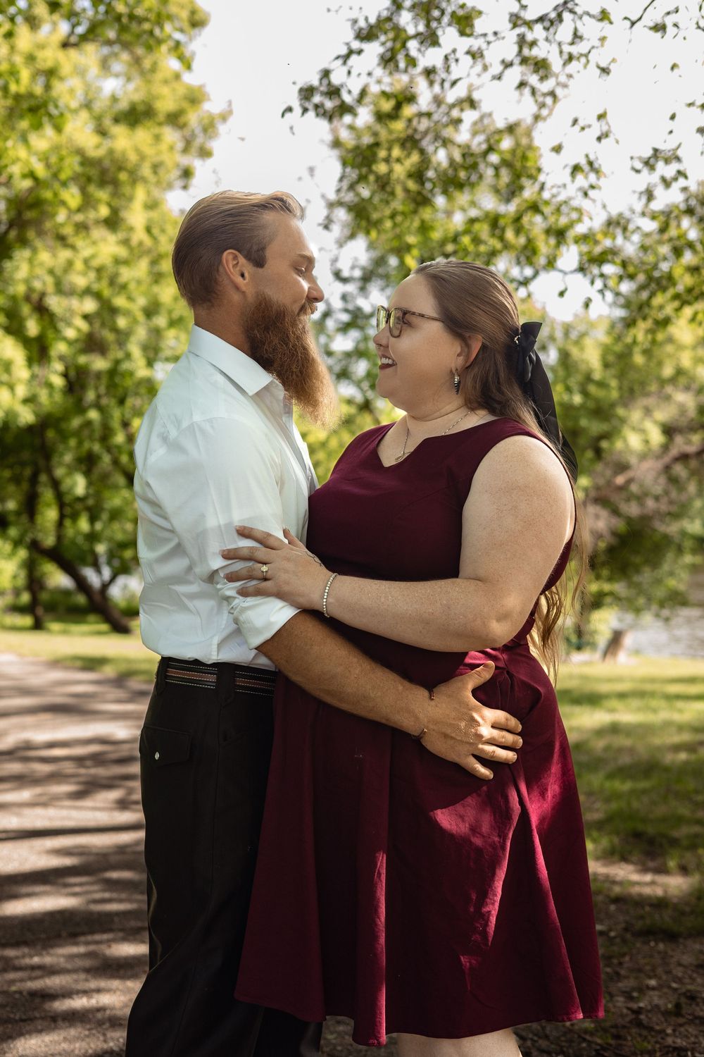 Smiling couple embracing in a sunny park, woman in burgundy dress and man in white shirt with red beard.