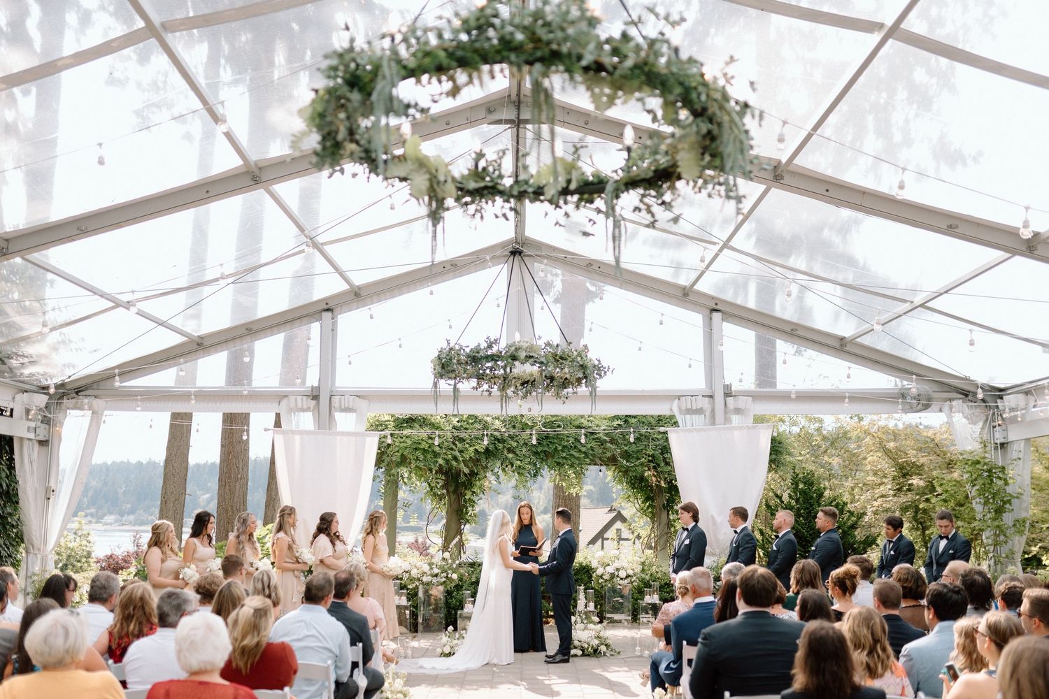 Wedding ceremony inside elegant clear tent with lush greenery chandeliers and guests watching couple at white altar.