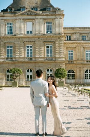 A couple stands outside an elegant French chateau with classical architecture and ornate windows on a sunny day. Paris film photoshoot.