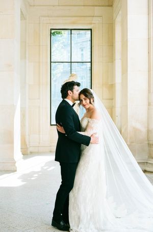 Bride in flowing white gown and groom in black suit share romantic moments in a sunlit colonnade hallway.