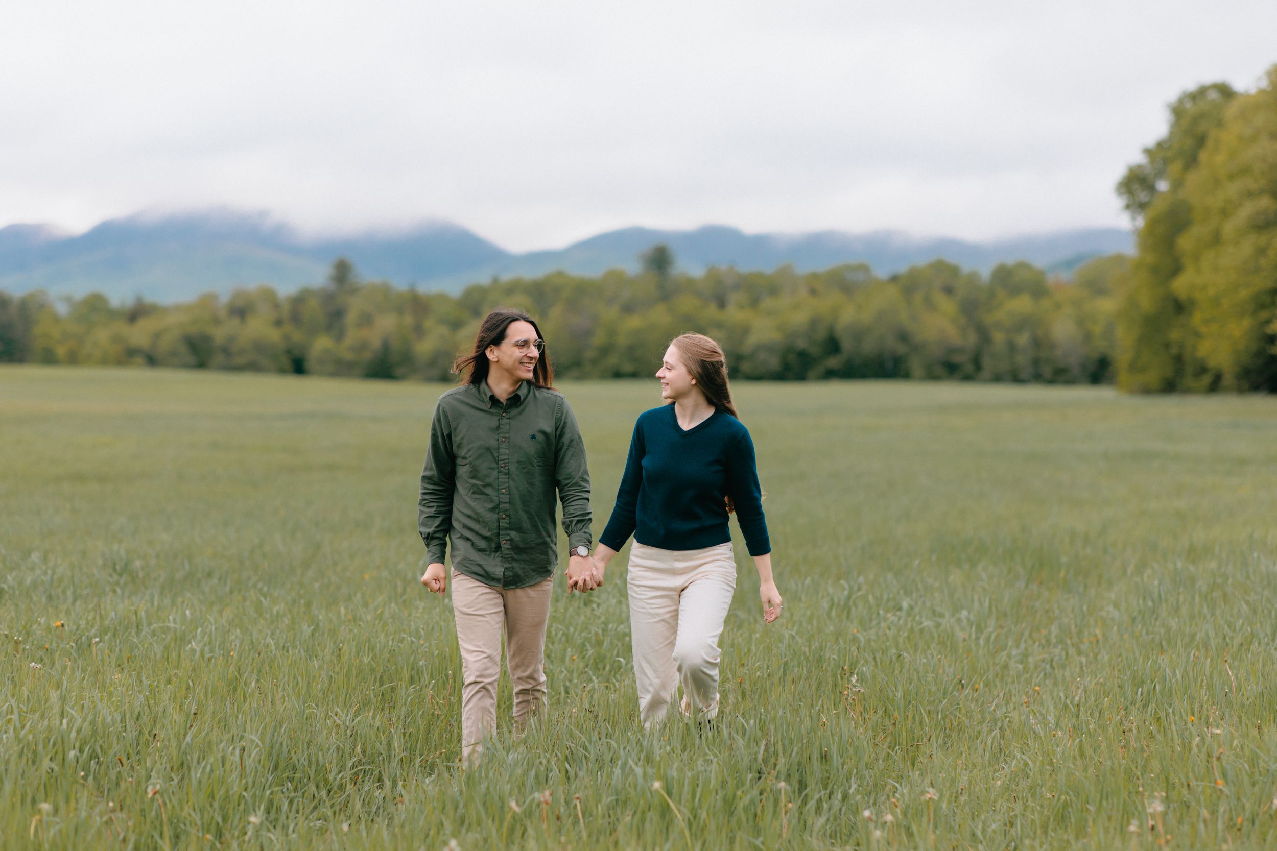 Two people hold hands while taking a stroll through a peaceful meadow with mountains and trees in the distance.