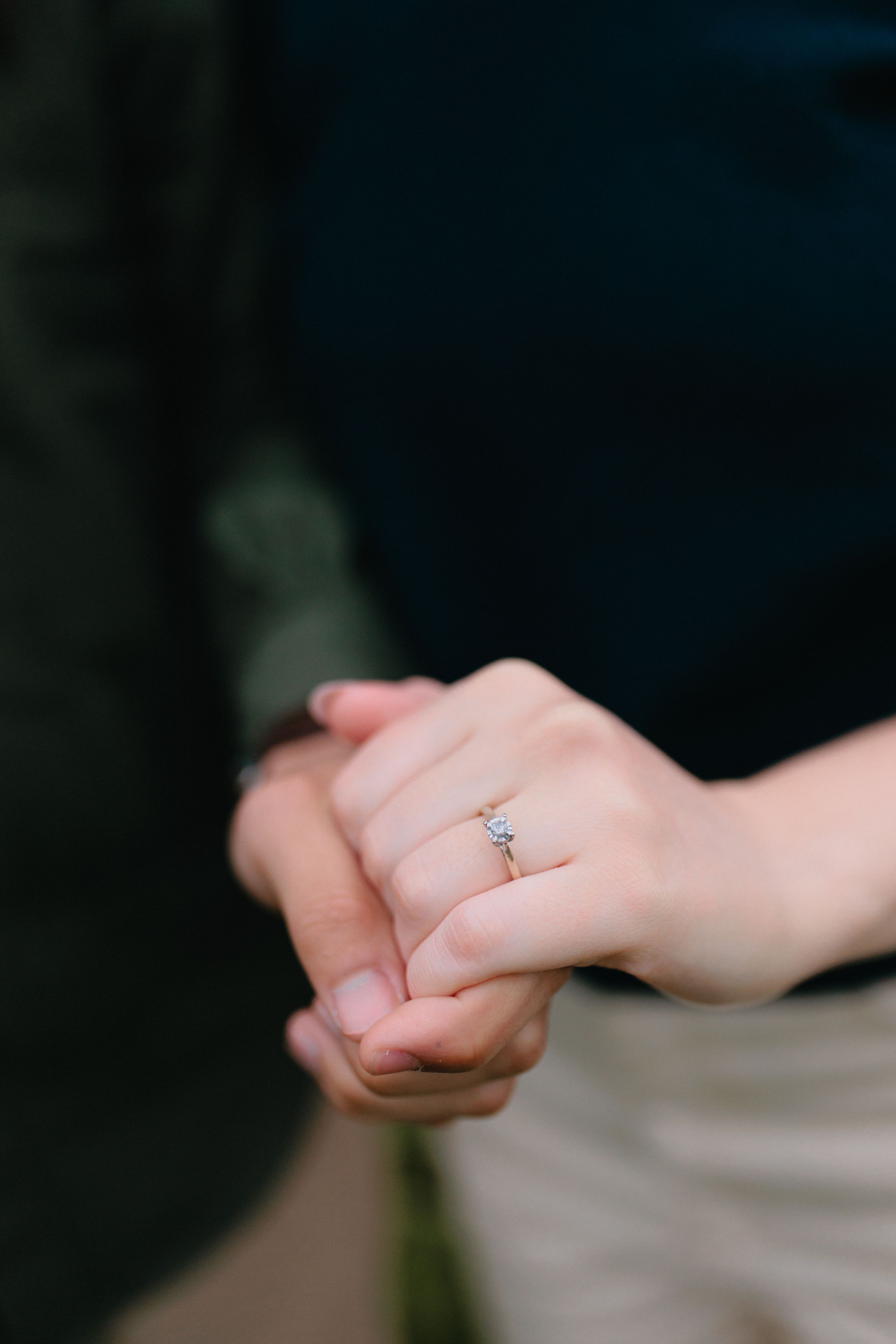 Close-up photo of clasped hands with delicate engagement ring featuring a small diamond on a simple band.