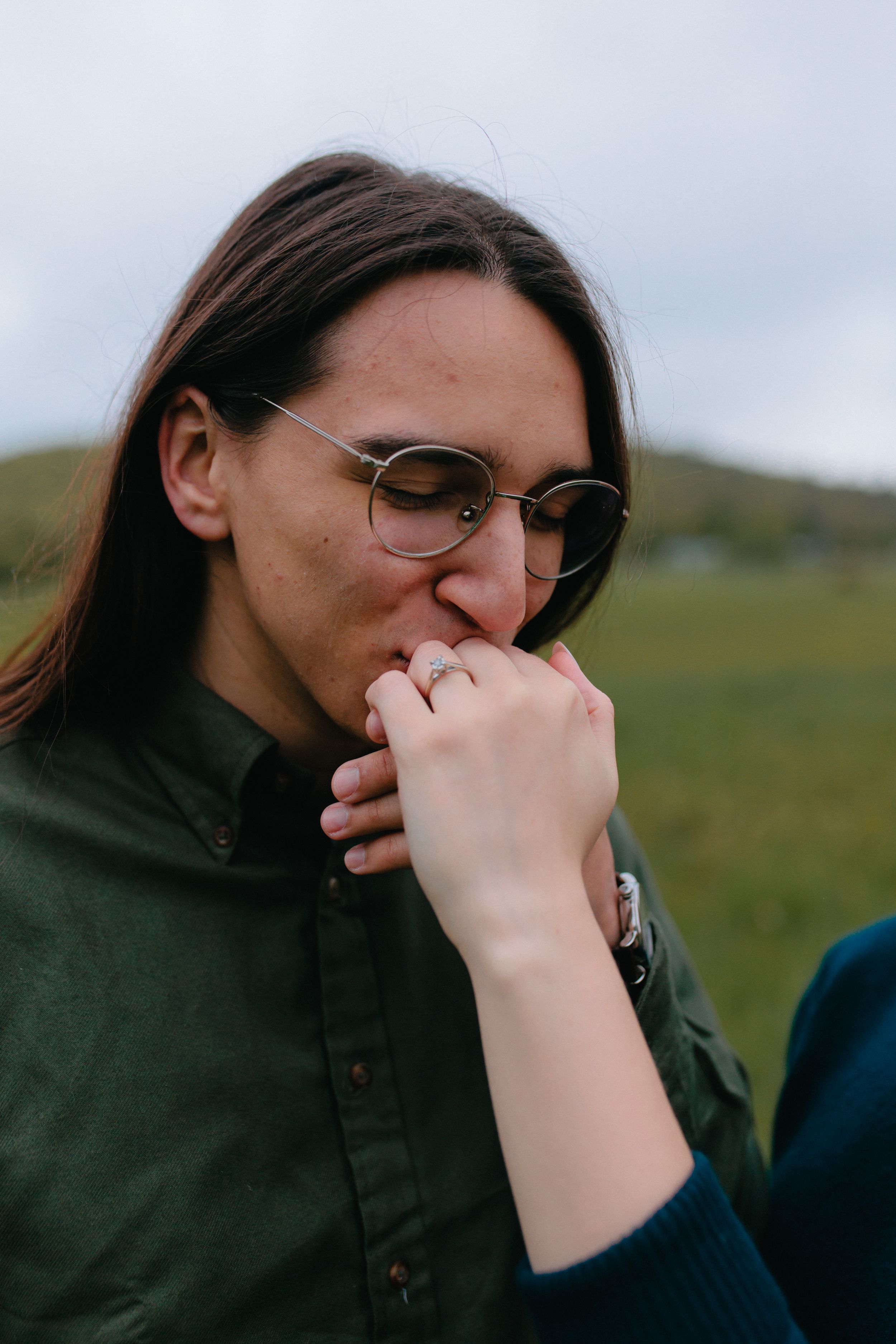 Person in a green jacket and glasses looks down thoughtfully in a natural outdoor setting.