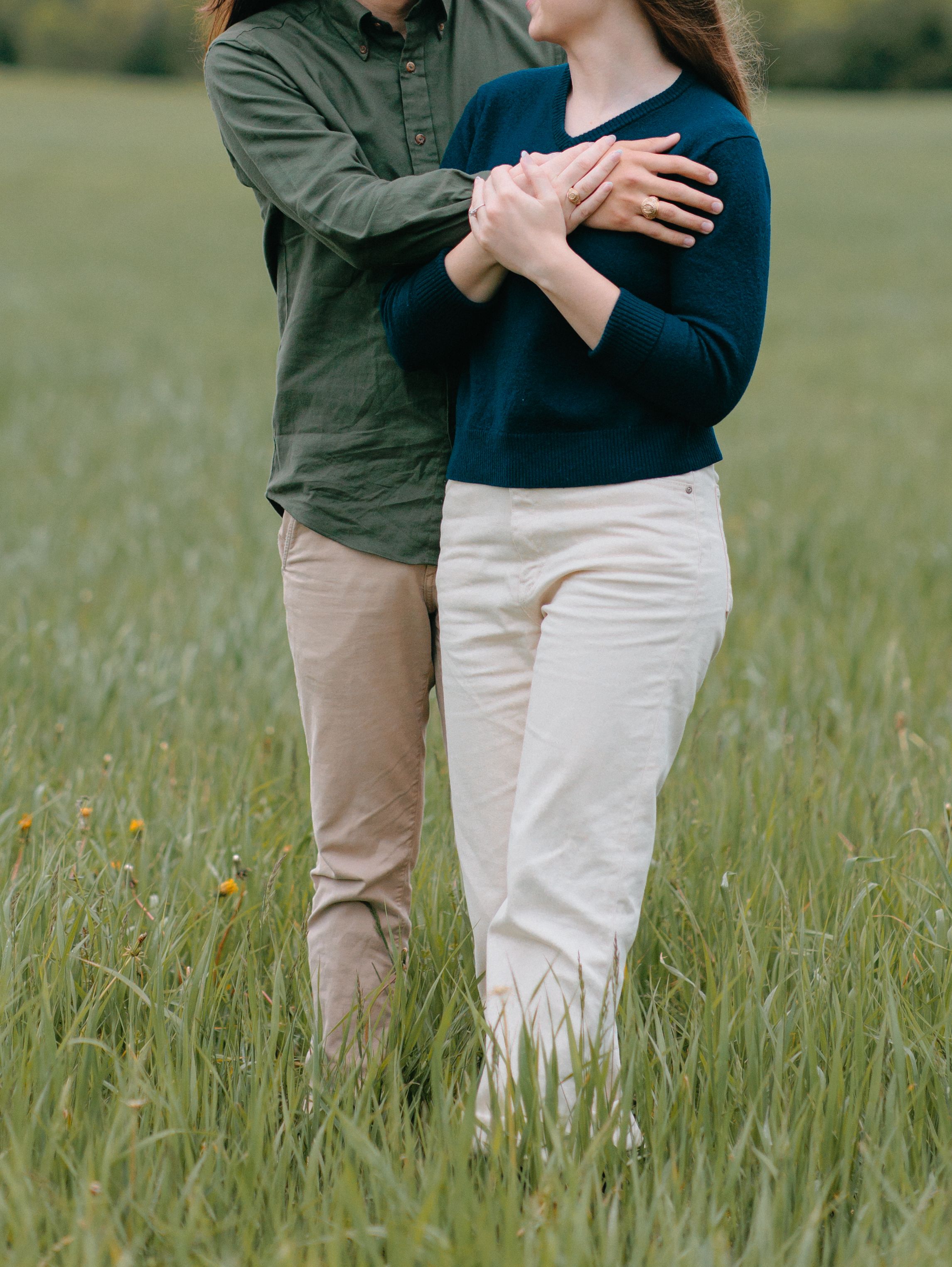 A couple in casual clothing stands close together holding hands in a green meadow filled with tall grass.