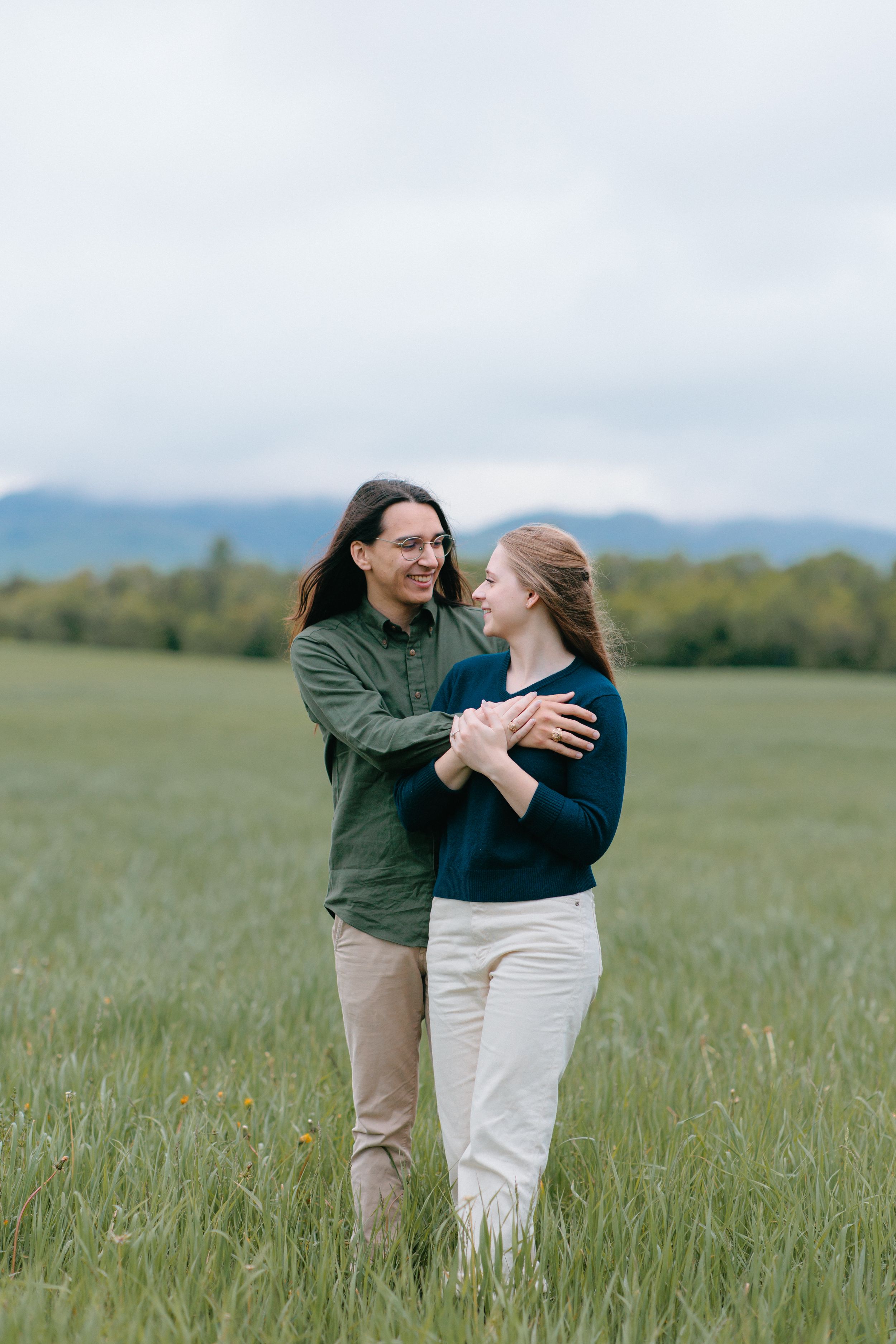 A couple shares a romantic embrace while standing in a meadow with mountains in the background.