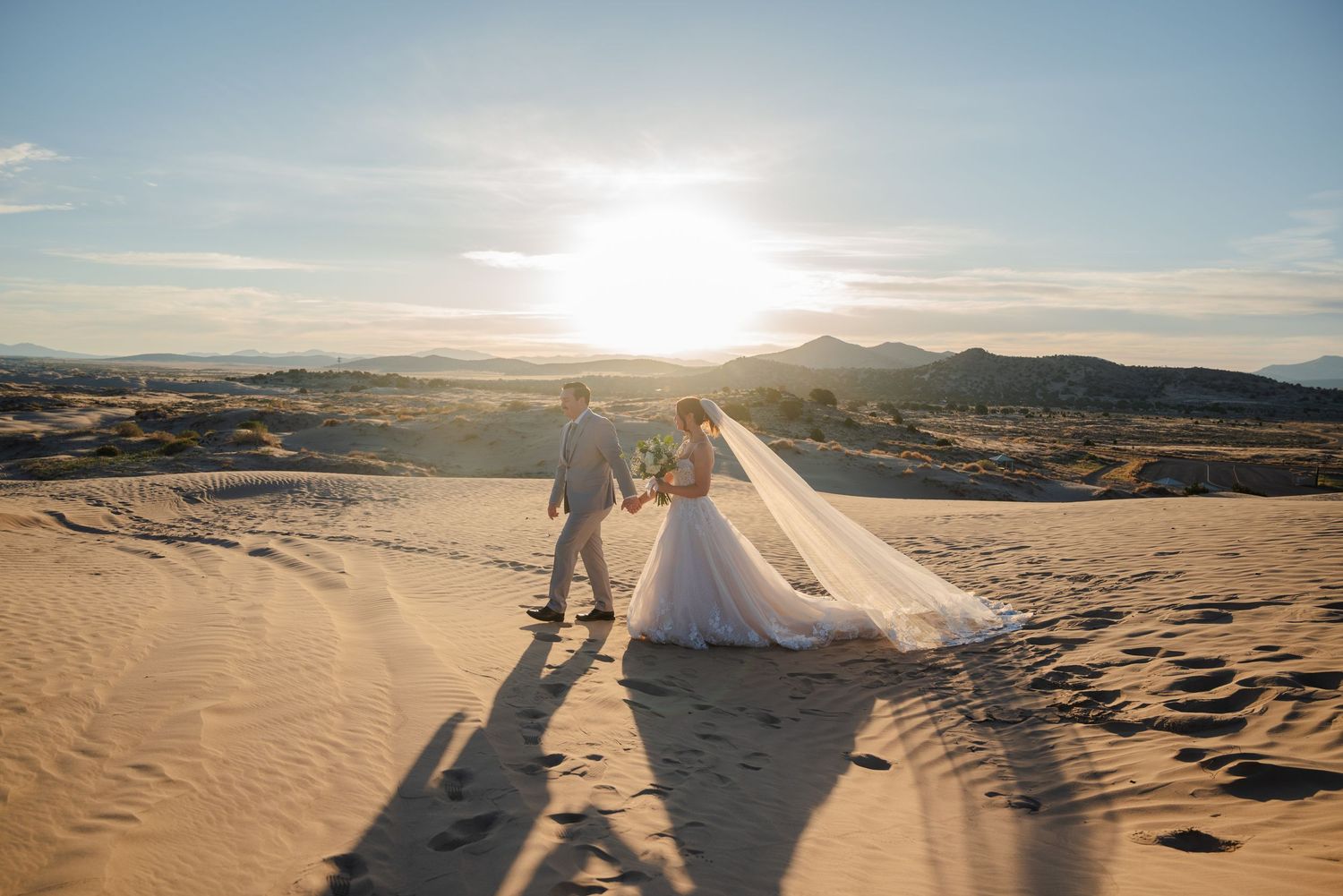 Bride and groom walk hand-in-hand across golden sand dunes at sunset with mountains in the background.