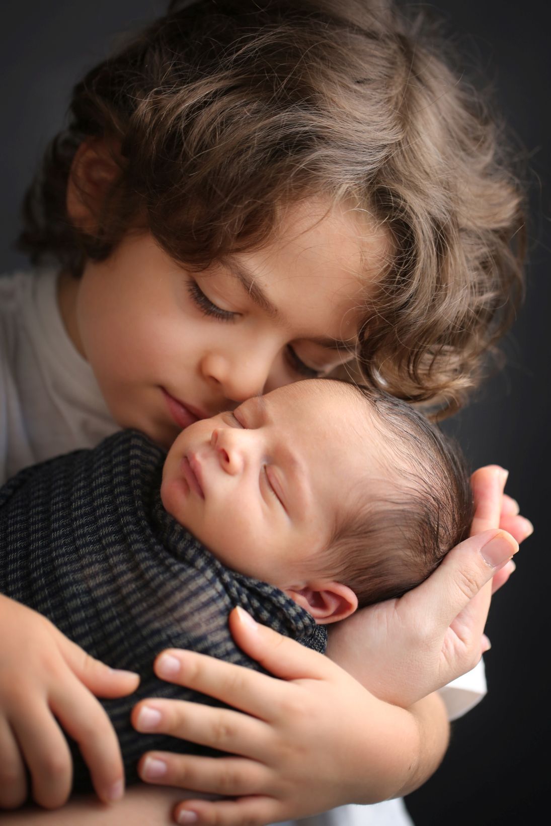 A tender moment shows older sibling gently hugging and kissing newborn baby who is wearing a striped top.
