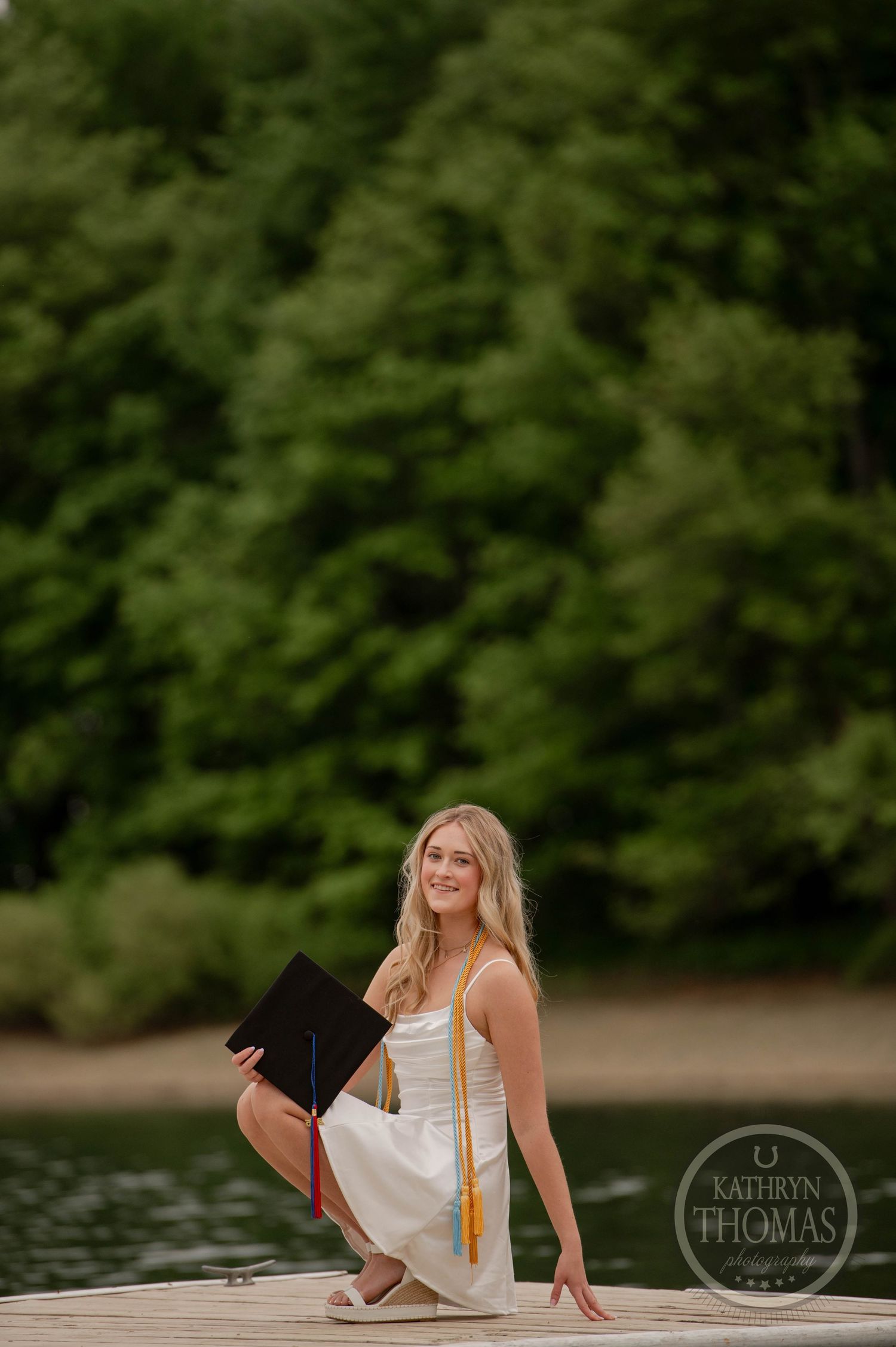 Graduate in a white dress sits on a wooden dock by a lake with green trees in the background during a photo session.
