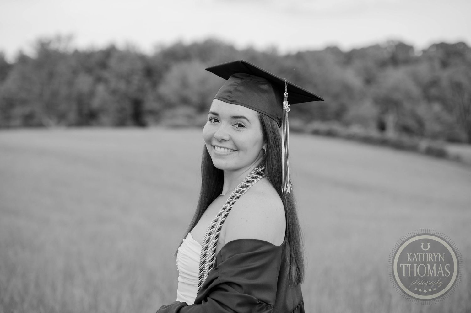 A graduate in cap and gown poses outdoors in a black and white portrait against a natural field backdrop.