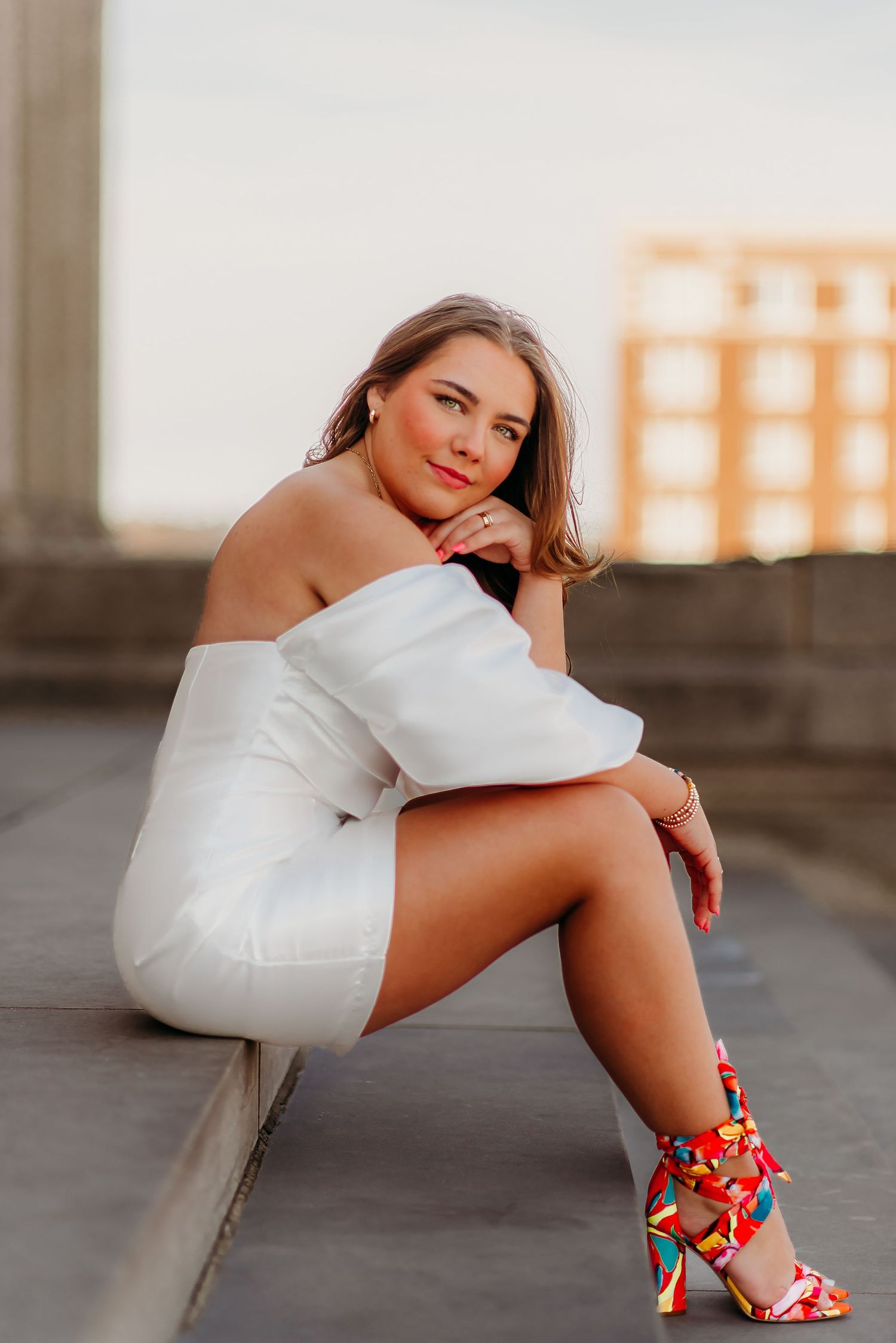Model poses outdoors in white off-shoulder dress and colorful floral heels during sunset golden hour.