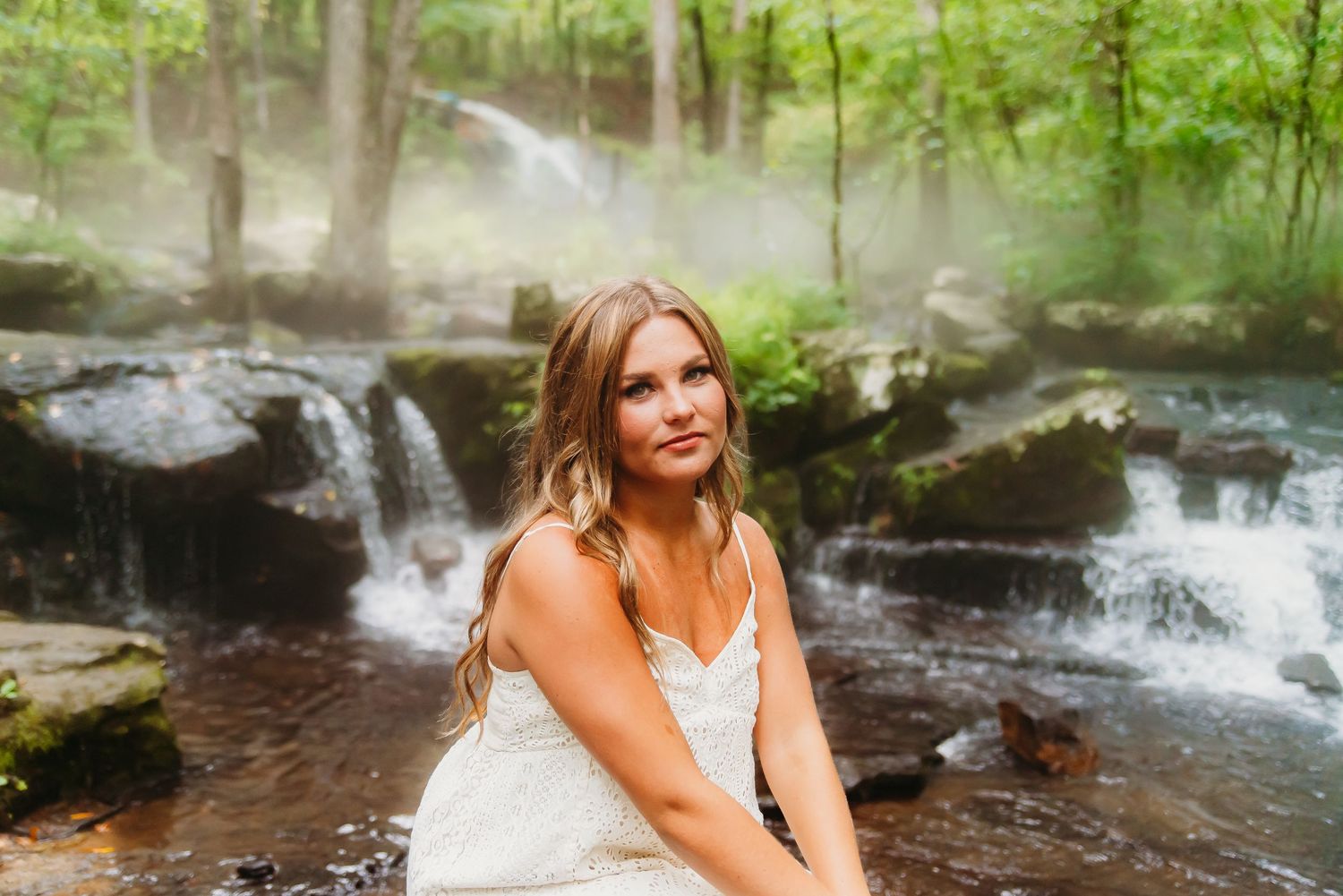 Person in white dress sits by cascading waterfalls in lush green forest setting.