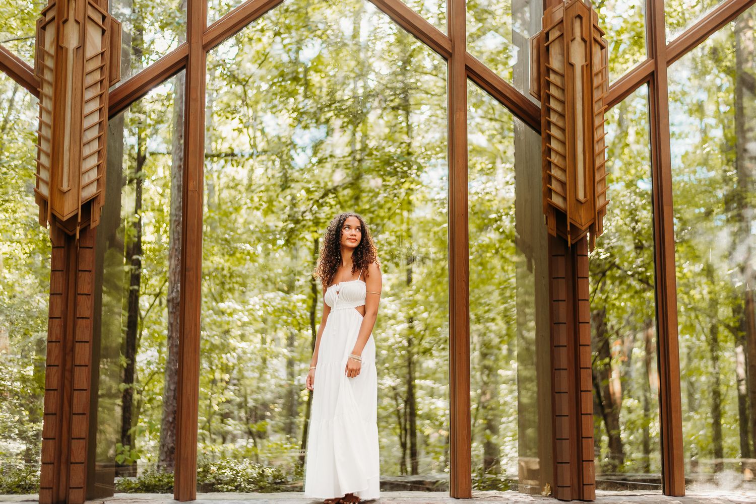 A person in a white dress stands between wooden architectural posts in a sunlit forest setting.