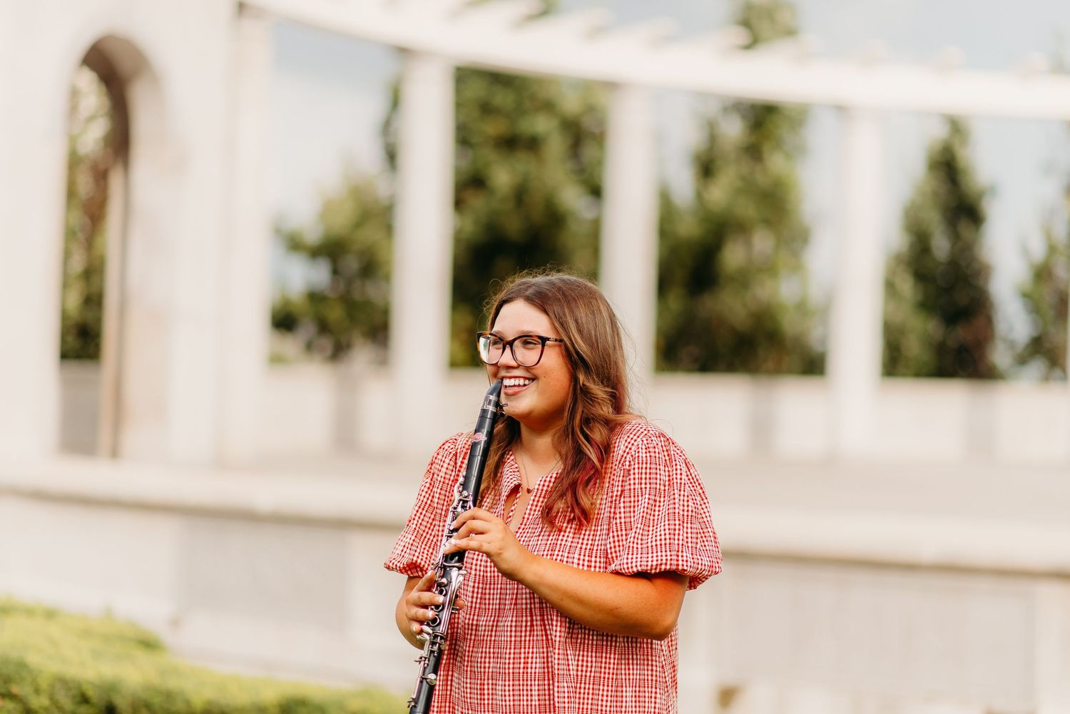 In front of white columns, someone in a pink striped dress stands and smiles while holding a drink outside.