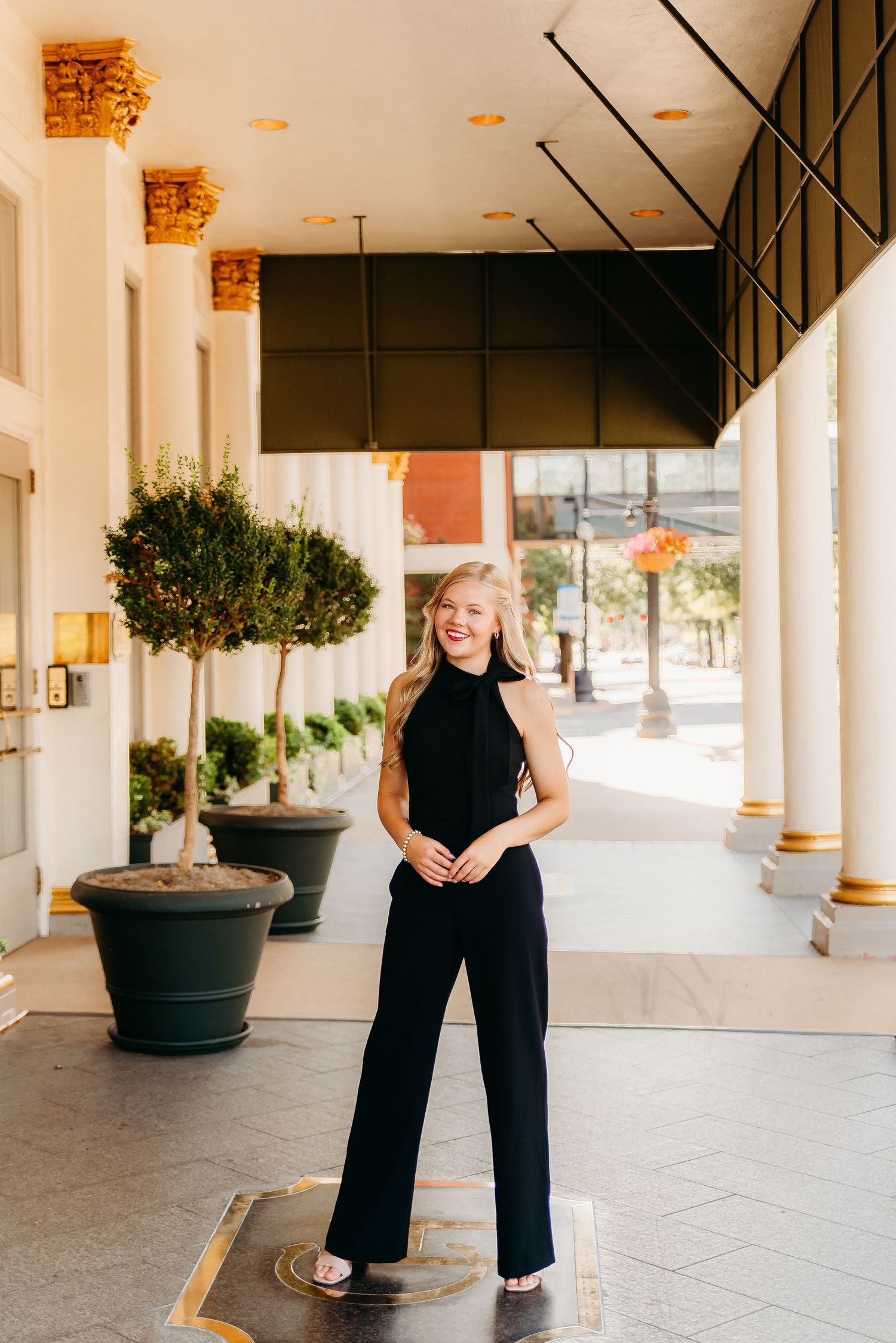 A person in a black outfit poses along a modern covered walkway with potted plants and columns.