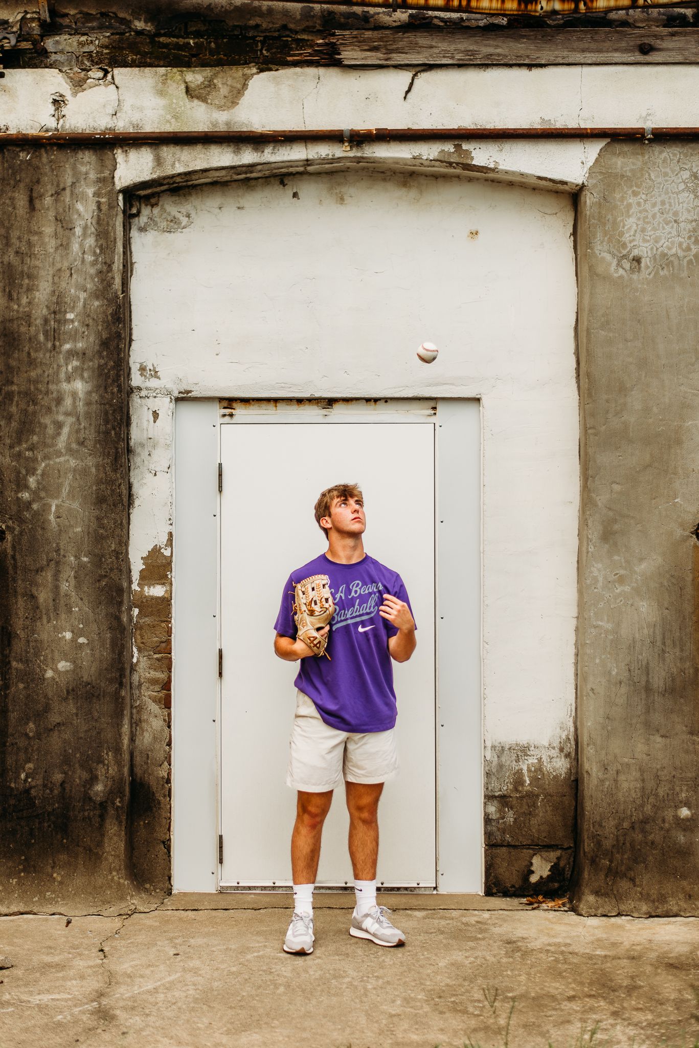 Person in purple t-shirt and white shorts stands against weathered white door with concrete wall backdrop.