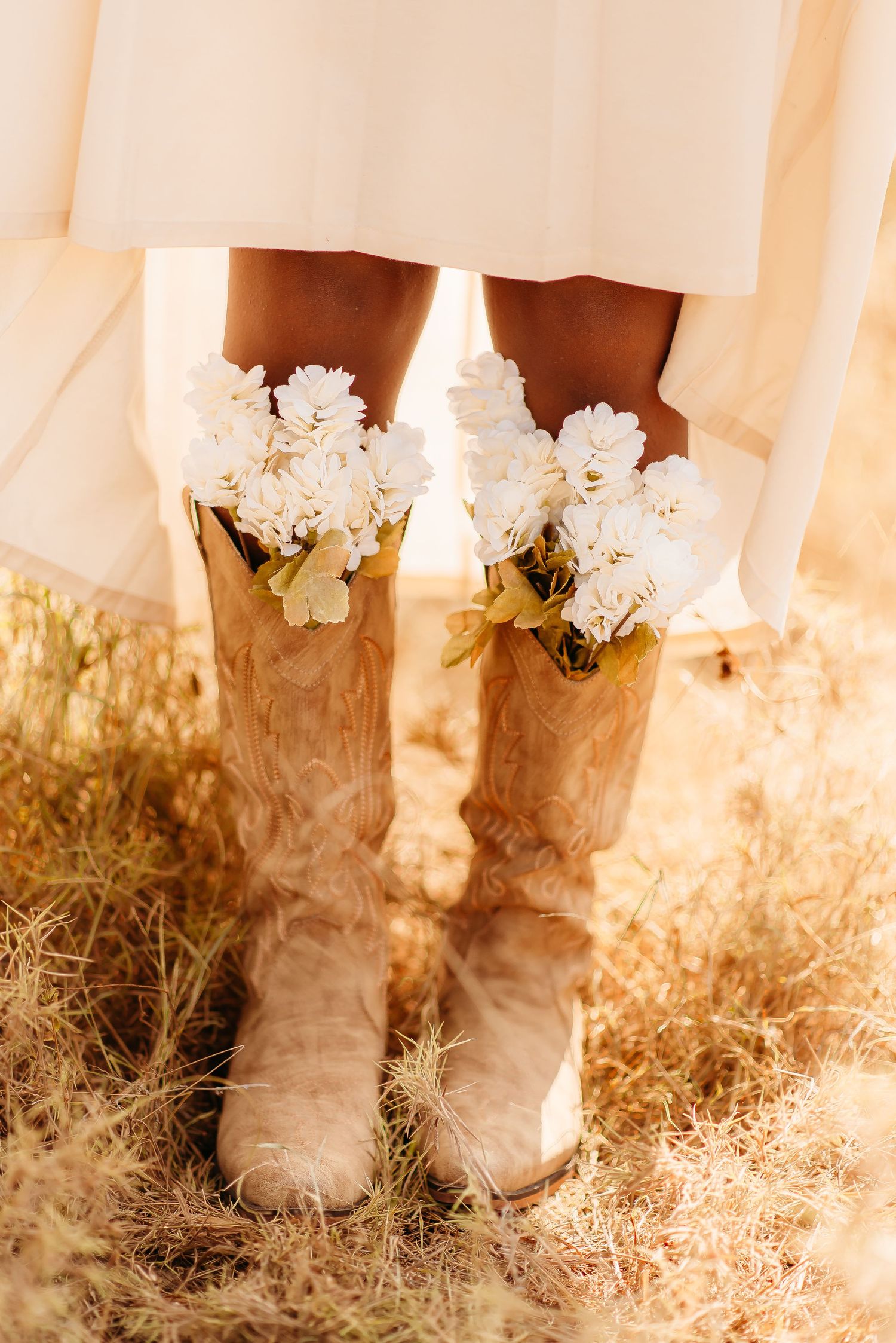 White cowboy boots decorated with delicate white flowers stand against a soft, dreamy background in golden sunlight.