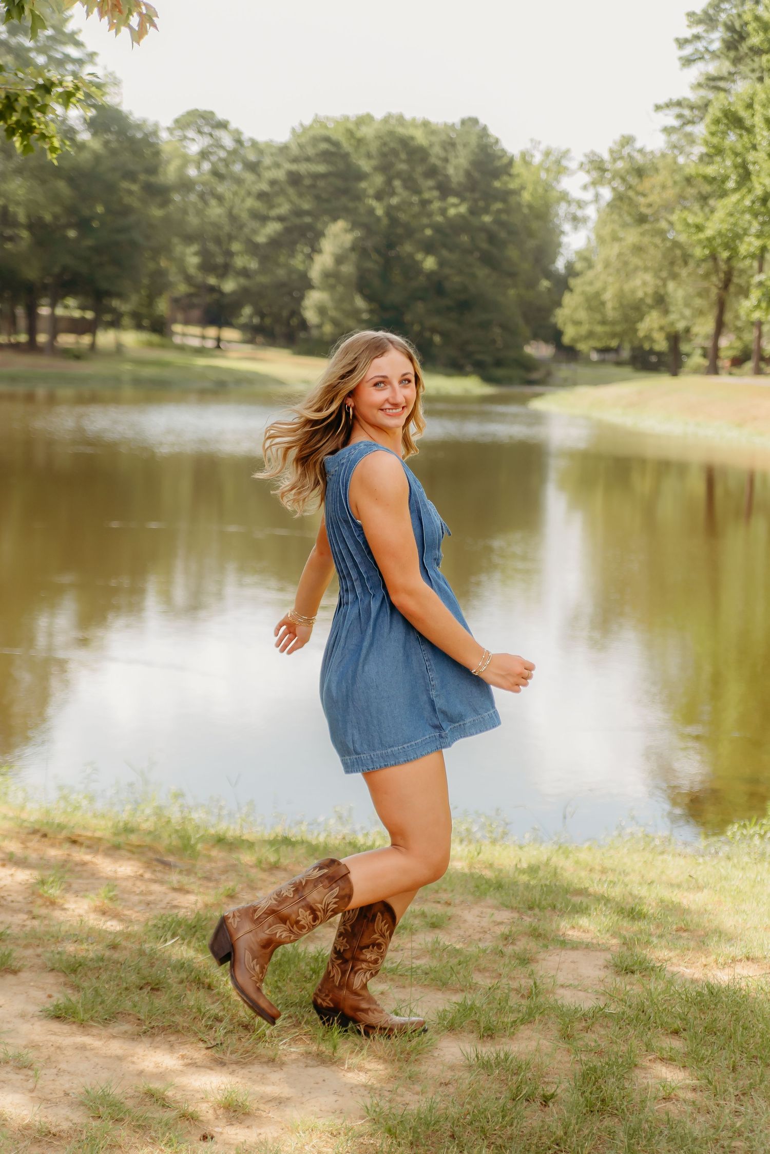 A person in a denim dress and brown cowboy boots poses playfully by a serene pond on a sunny summer day.