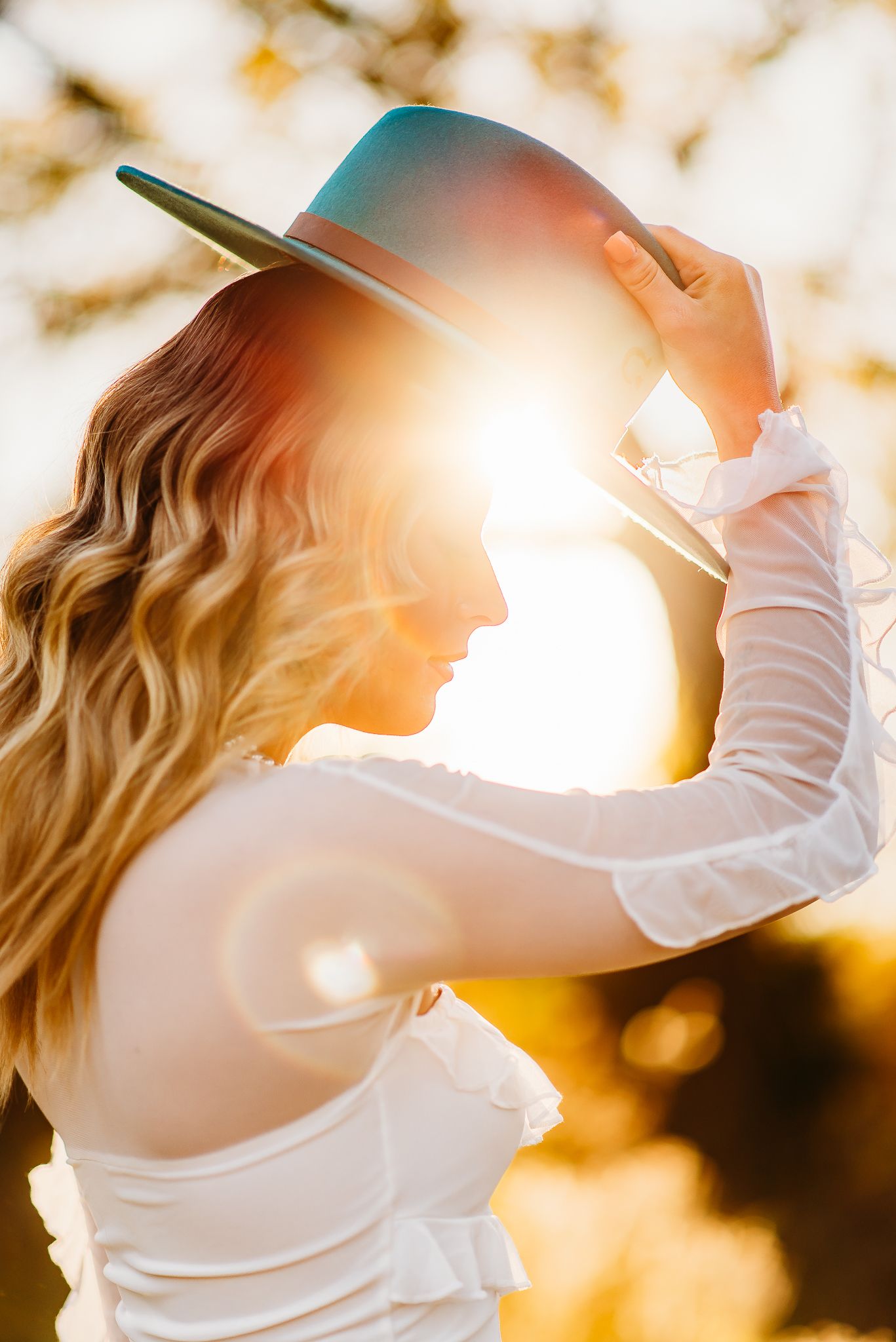 A woman in a white dress and hat poses in silhouette against golden sunlight during a sunset photoshoot.