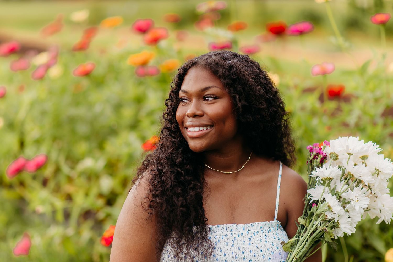 A natural curly-haired person in a white top smiles warmly in a garden with red and white flowers.