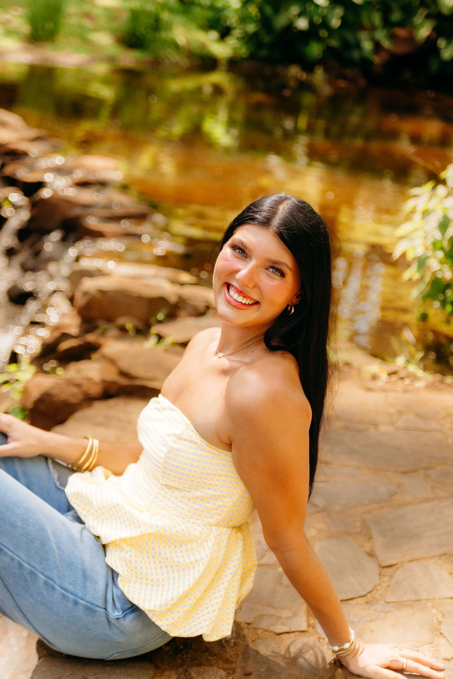 A casual outdoor portrait captured by a creek with rocks, featuring yellow top and denim in warm natural lighting.