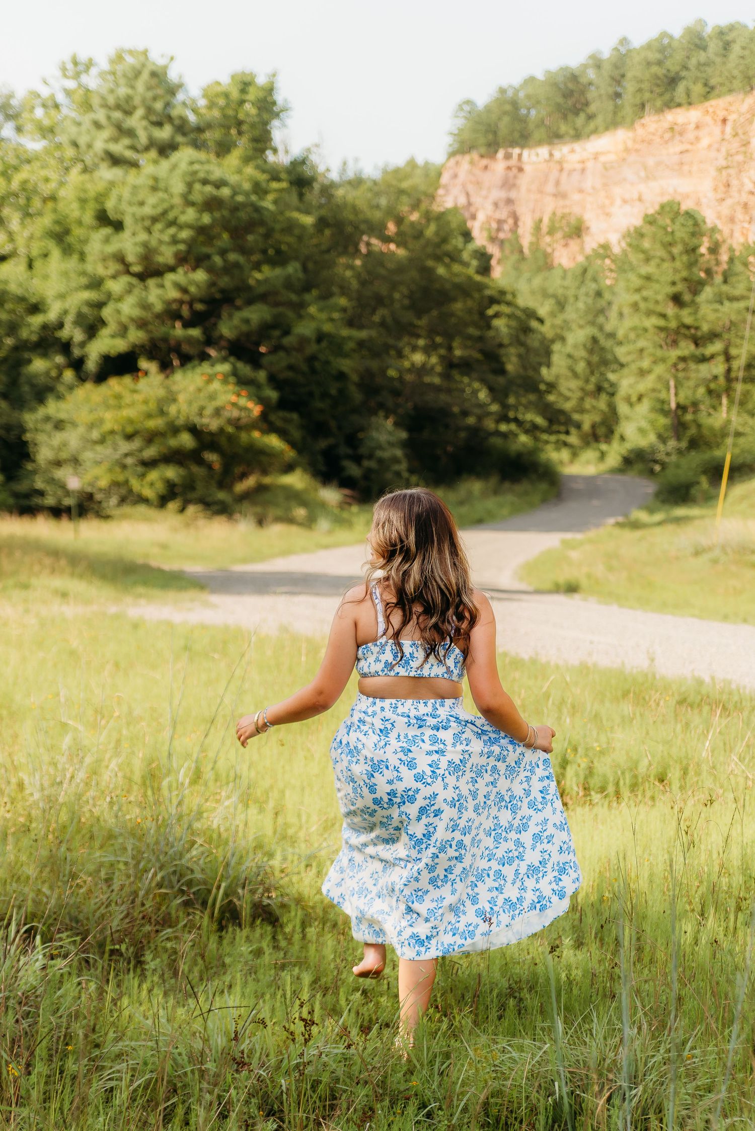 Multiple images show a person in a floral blue dress twirling and dancing in a sunlit meadow with trees and cliffs in background.
