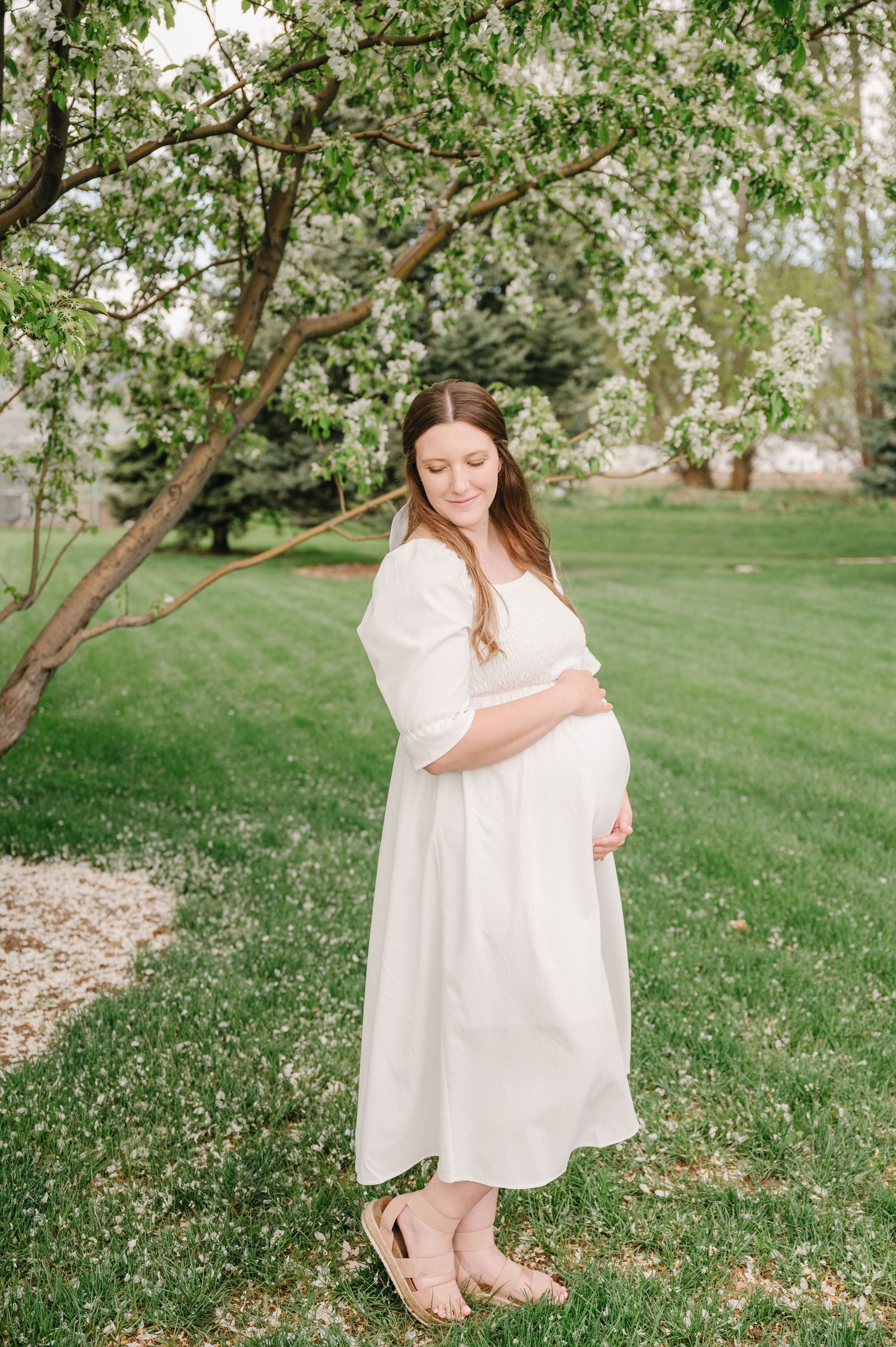 An expectant mother in a flowing white dress stands serenely under a blooming spring tree in a lush garden setting.