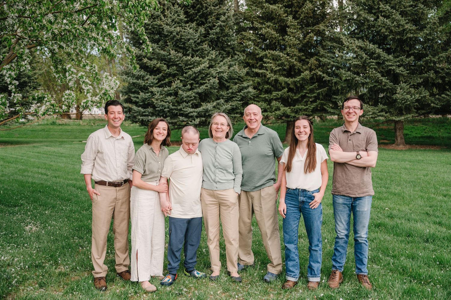 A group of seven people standing together on a green lawn with evergreen trees in the background.