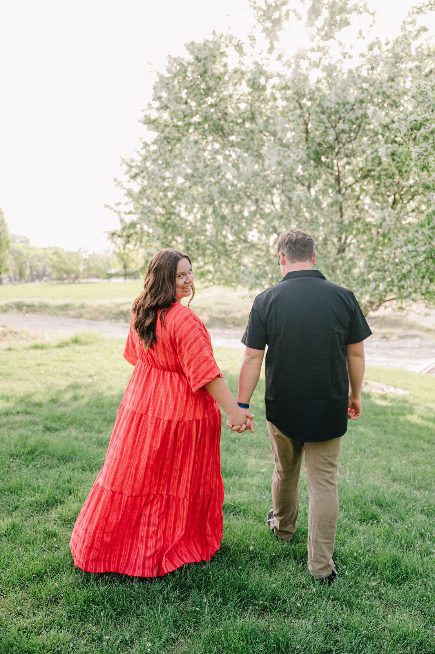 Two people holding hands while walking on green grass with blooming trees in the background on a sunny day.