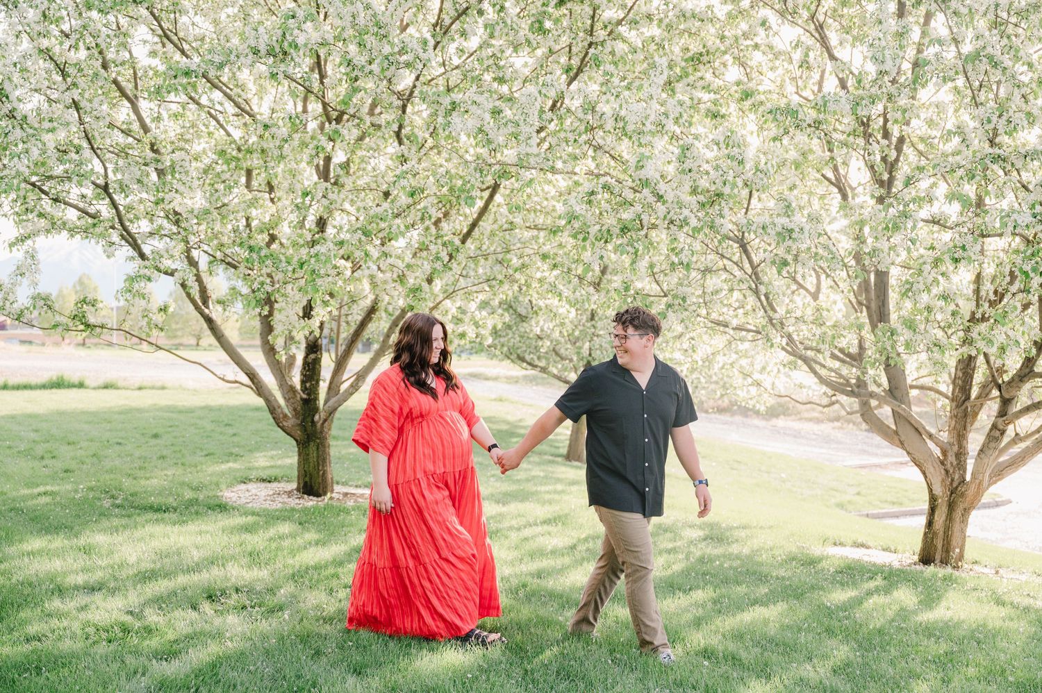 A couple walks together through a blooming spring orchard with a person wearing a flowing red dress and black shirt.