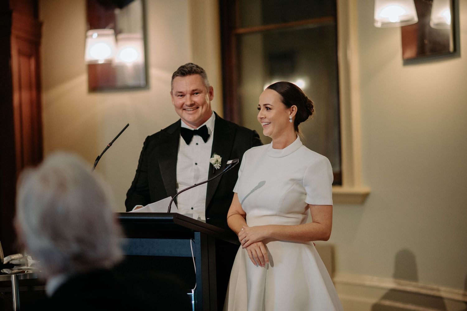 A couple stands at a podium together during their wedding reception, with the bride wearing a classic white dress.