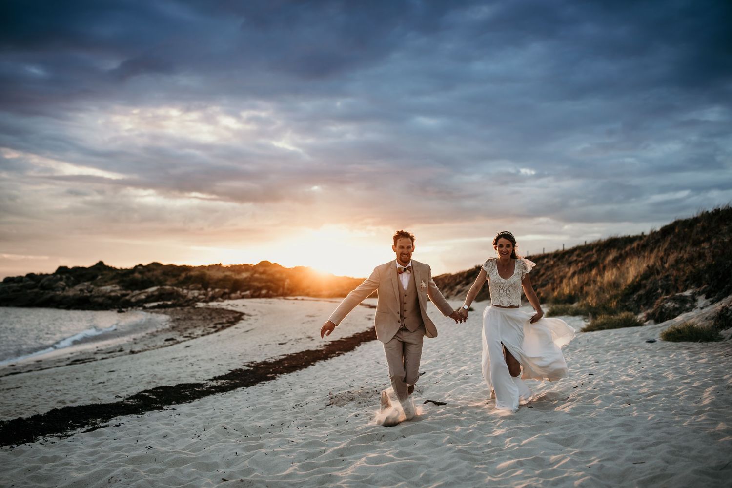 Couple walking hand in hand on beach with dramatic sunset and cloudy sky.