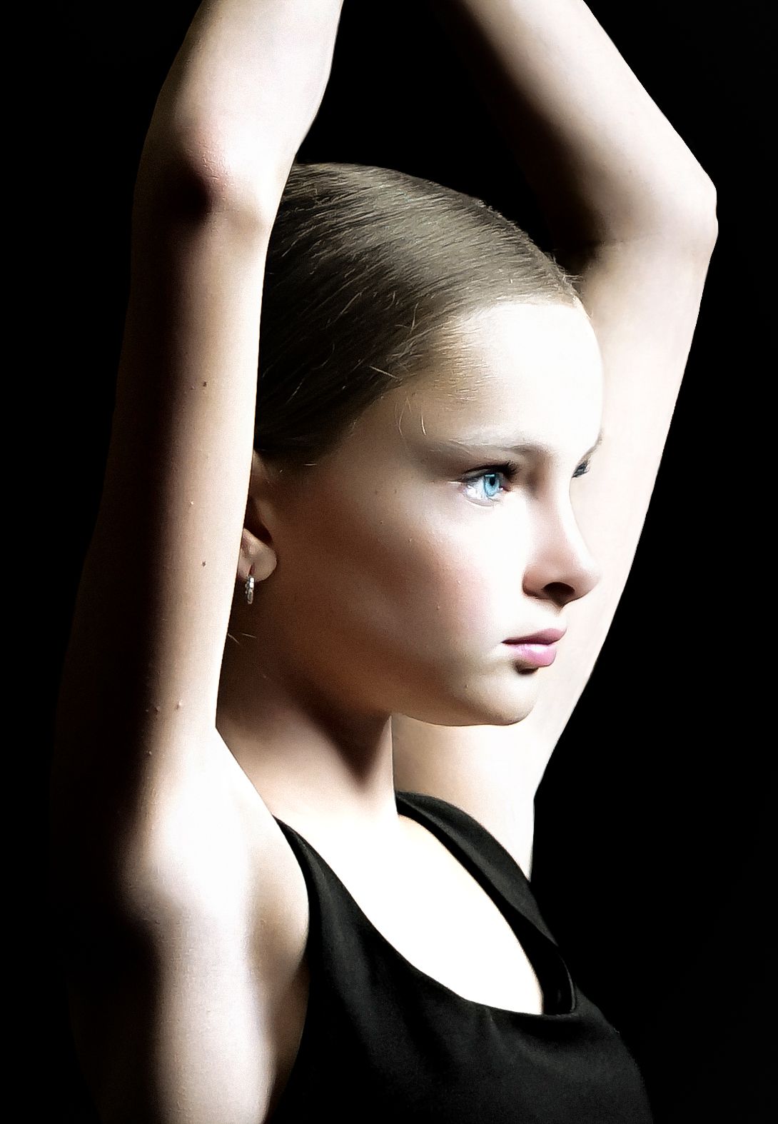 Young female dancer in black top raises arms gracefully against dark background, showcasing elegant ballet pose.