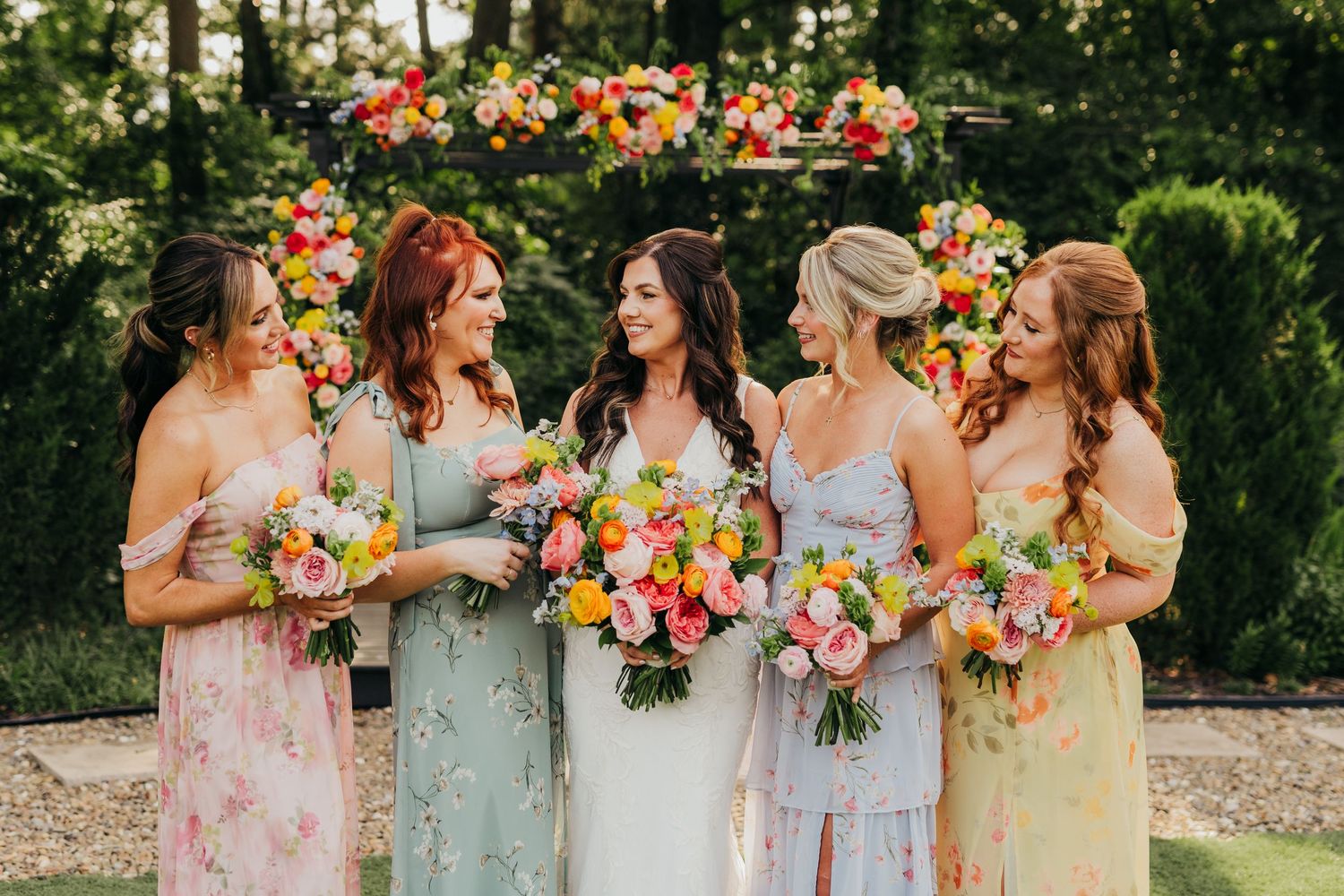 Bridesmaids wearing pastel dresses stand together holding vibrant bouquets of flowers in a garden setting in East Texas.