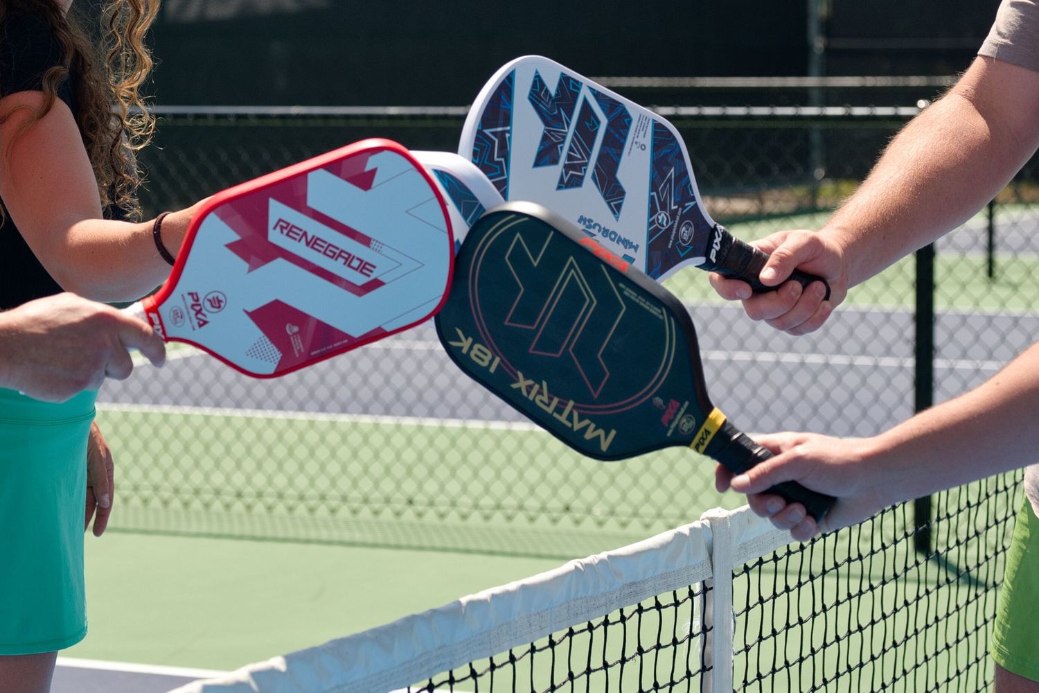 Pickleball players tap paddles together over the net on an outdoor court in a friendly gesture of sportsmanship.
