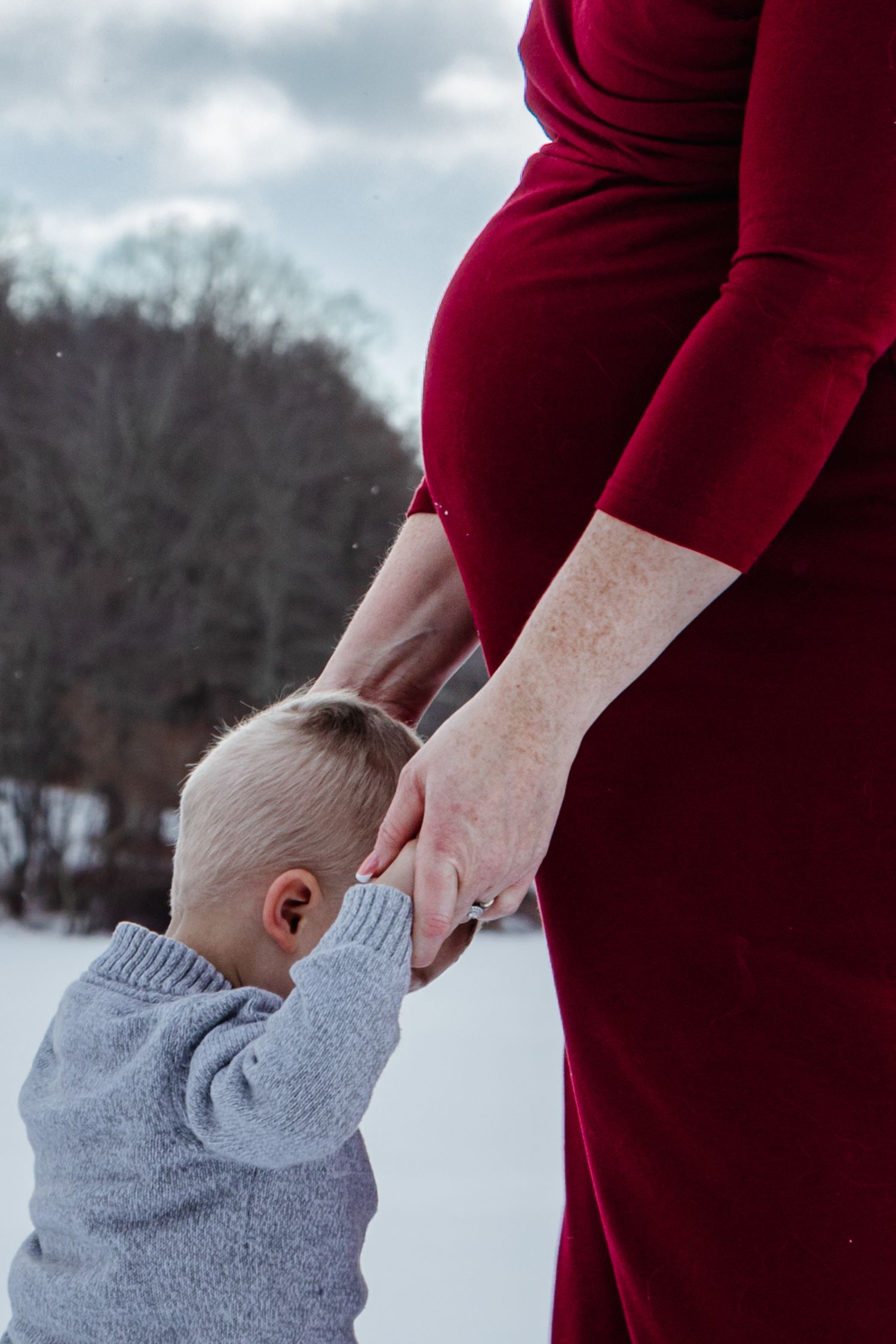 Close-up of a toddler holding the hands of his pregnant mother in a red dress, standing together in a snowy winter landscape