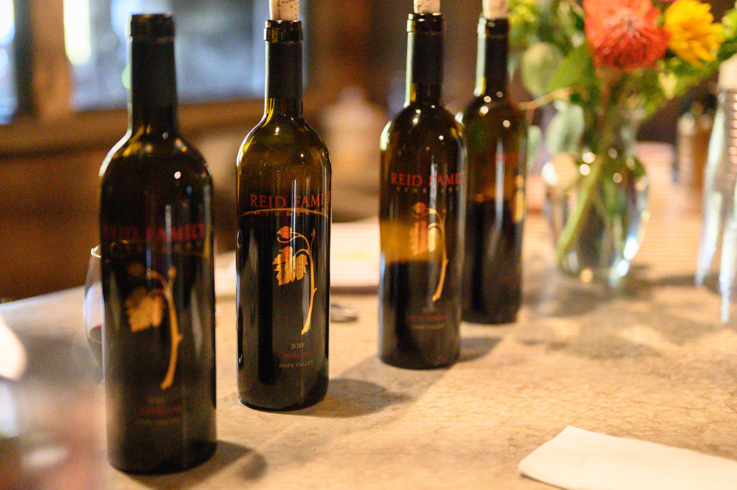 Row of dark wine bottles arranged on a rustic table with colorful flowers in a vase at the far end.