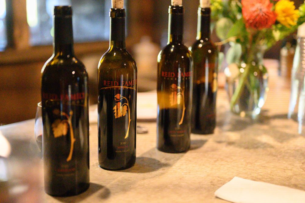 Row of dark wine bottles arranged on a rustic table with colorful flowers in a vase at the far end.